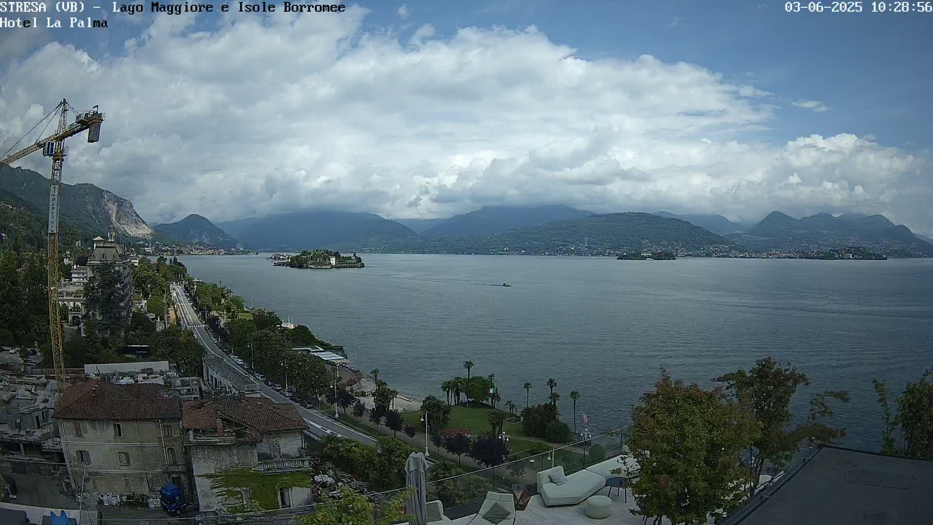 A partly cloudy day overlooks a lake with islands and mountains in the distance, a city and roadway below, and a crane to the left.