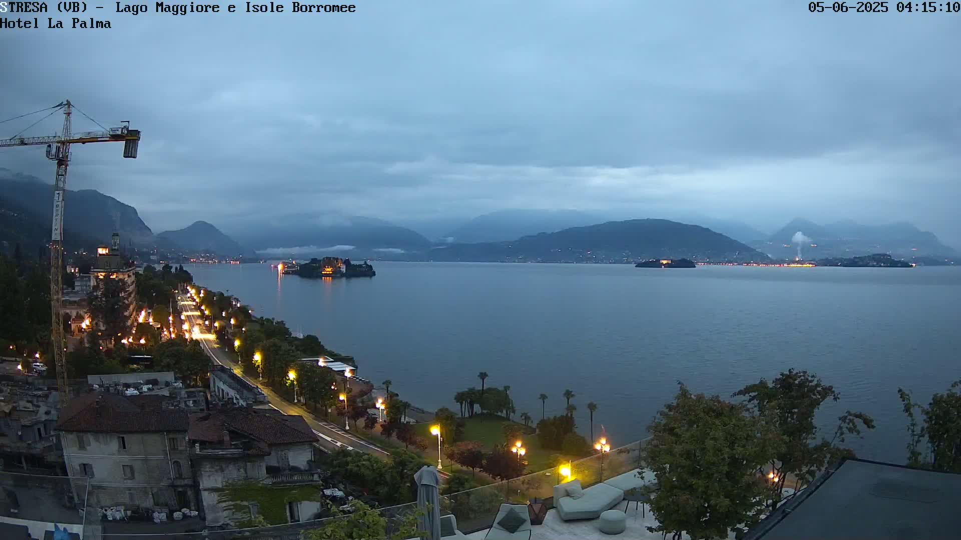 A twilight view of a calm lake with islands and mountains in the background, a road with lights along the shoreline, and a crane visible on the left.
