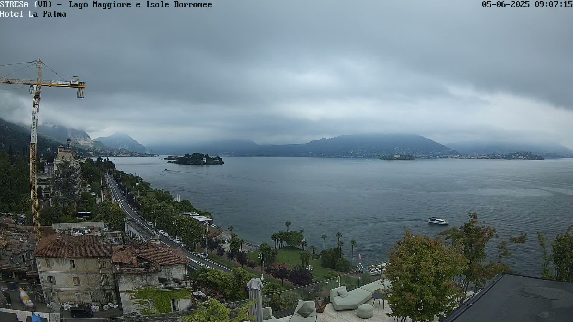 A view of a lake with islands and mountains in the background under an overcast sky, seen from an elevated vantage point overlooking a town with a construction crane visible.