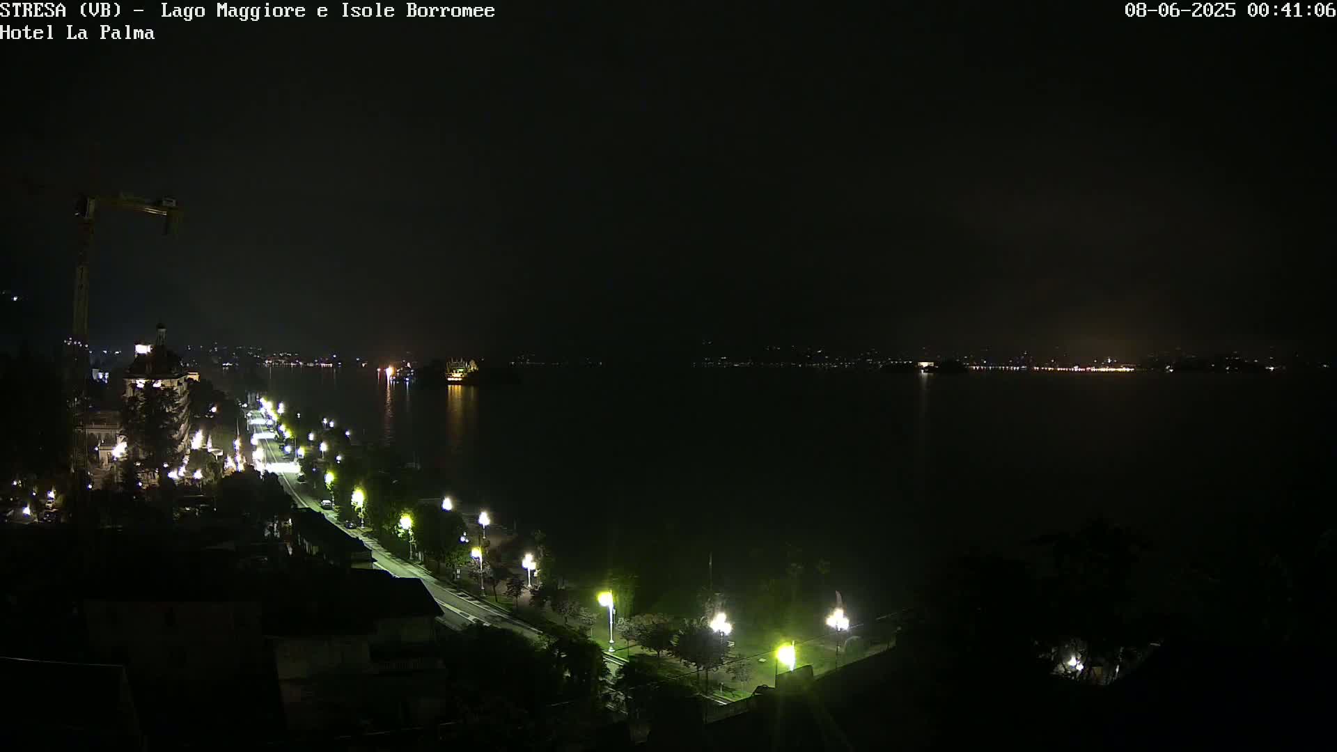 A nighttime view of a dark body of water with a brightly lit waterfront road and buildings along one shore, and a distant city skyline visible across the water.