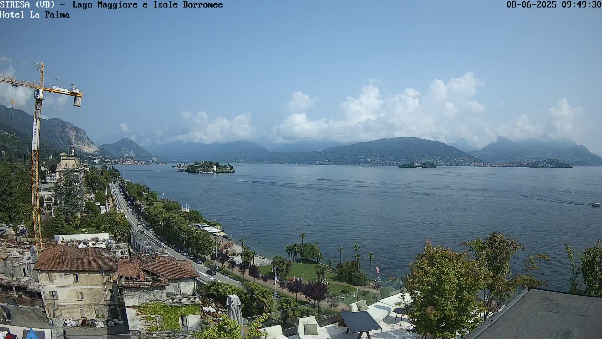 A partly cloudy day reveals a lake with islands and mountains in the distance, viewed from a high vantage point overlooking a town with buildings and a road.