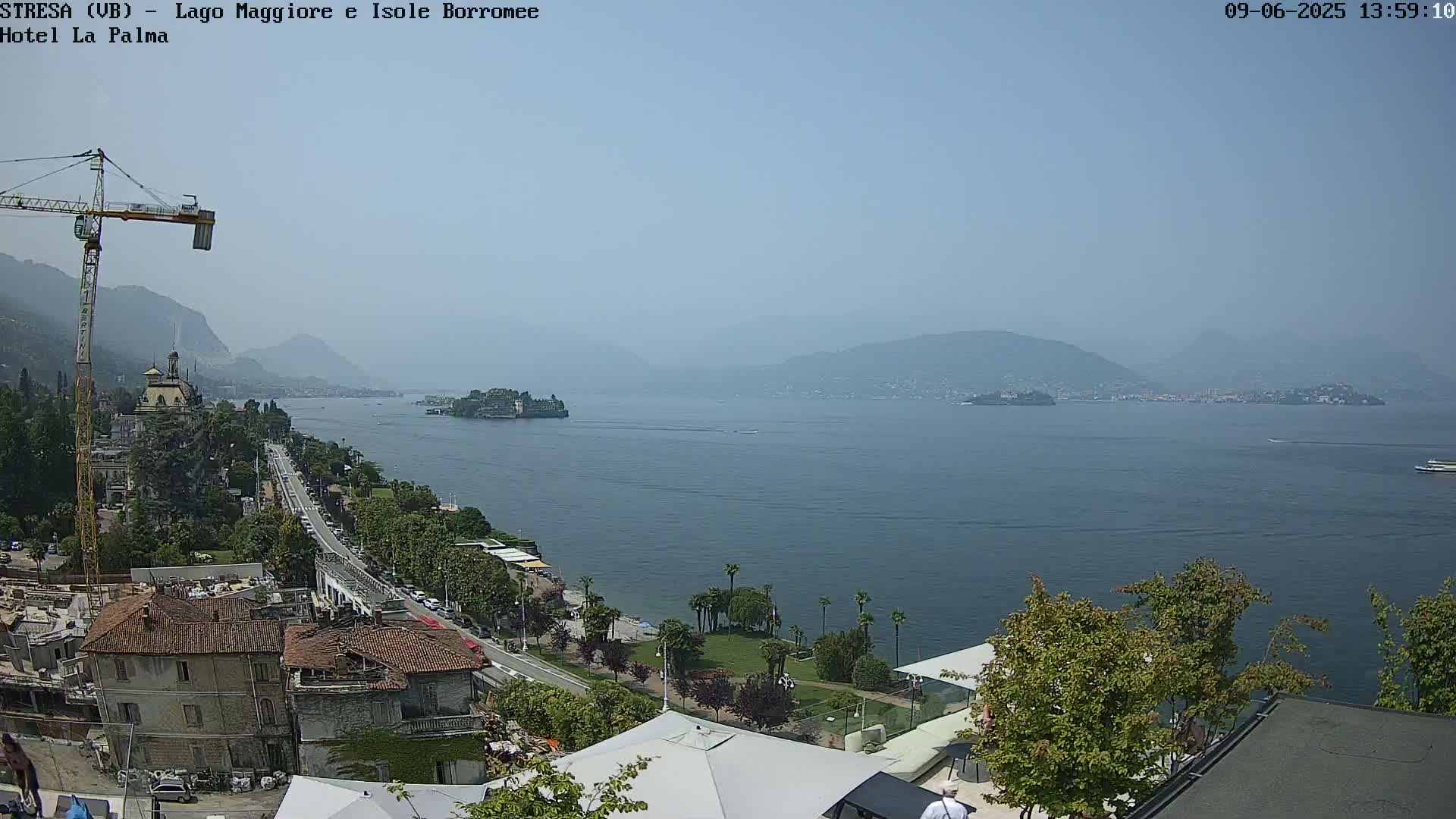 A hazy, sunny day overlooks a calm lake with a town, buildings, and a construction crane on the shore, distant islands, and mountains in the background.
