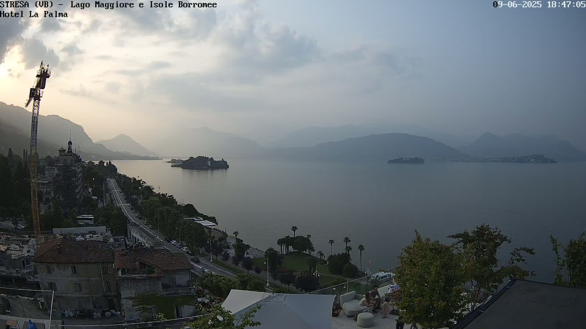 A hazy, late afternoon view of a calm lake surrounded by mountains and a lakeside town with buildings and palm trees, featuring a crane visible in the foreground.