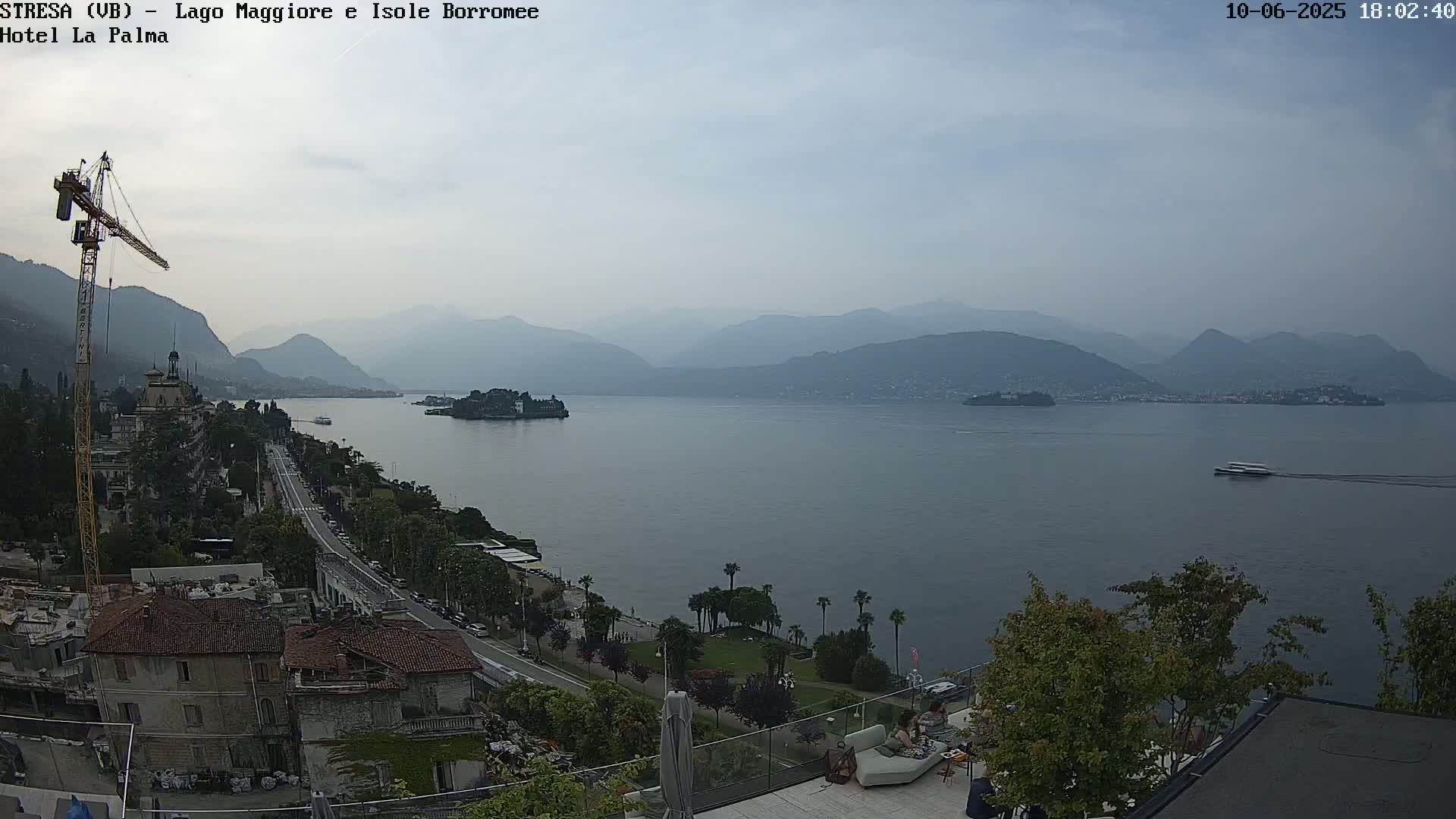A hazy, overcast view shows a large body of water with islands in the distance, mountains in the background, and a town with buildings and a construction crane in the foreground.