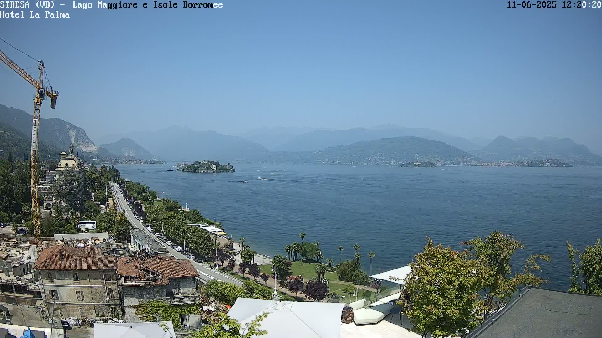 A clear, sunny day overlooking a large lake with mountains in the background, featuring a town with buildings and a construction crane along the shoreline.