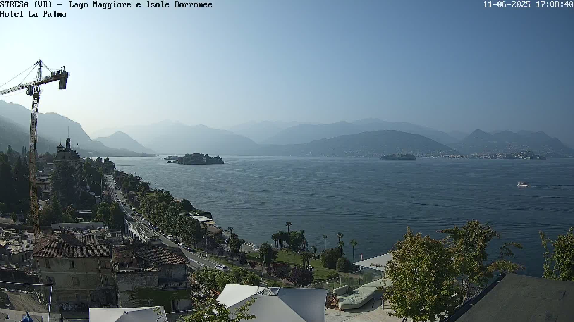 A clear, sunny day overlooking a large lake with mountains in the background, a town along the shore, and a construction crane visible in the foreground.