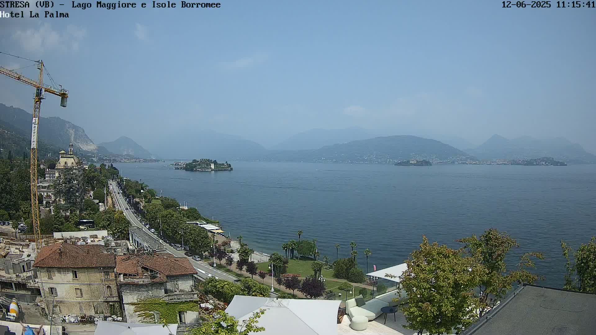 A sunny view of a lake and distant mountains, with a shoreline featuring buildings, trees, and a road.