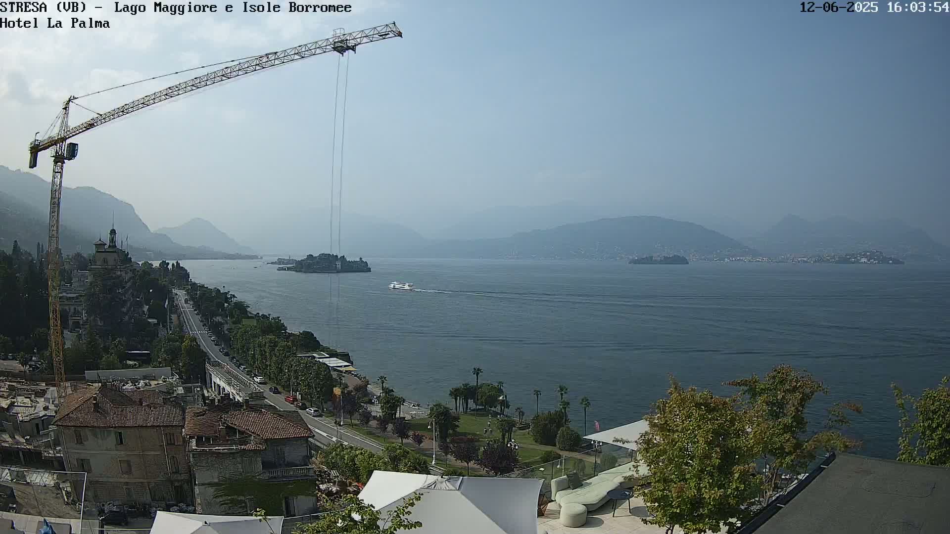 A large construction crane overlooks a hazy, sunny lake with islands and mountains in the distance, and a town with buildings in the foreground.