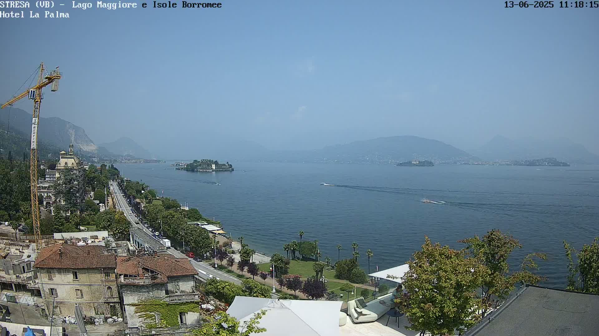 A sunny day overlooking a calm lake with buildings, trees, and a construction crane along the shoreline, and distant mountains in the background.