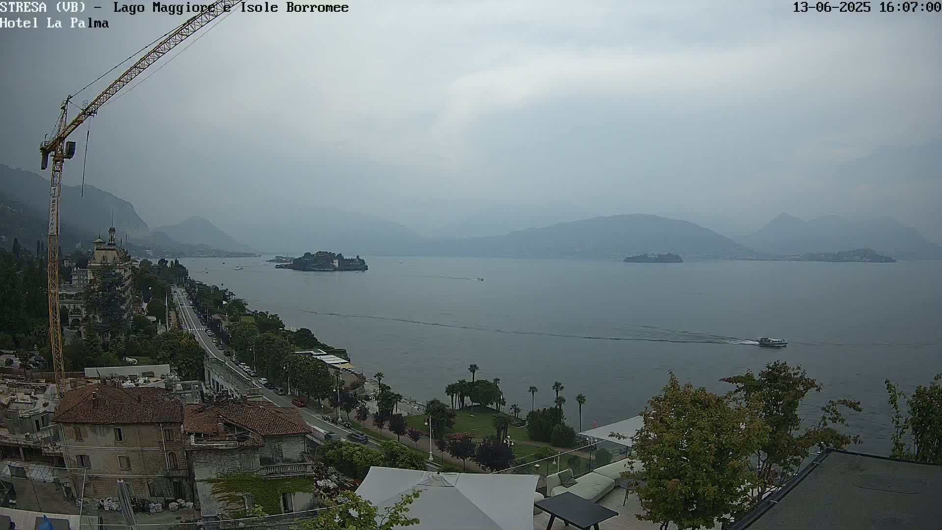 A hazy view of a lake with several islands and distant mountains, a shoreline town with buildings and a road, and a construction crane is visible on the left.