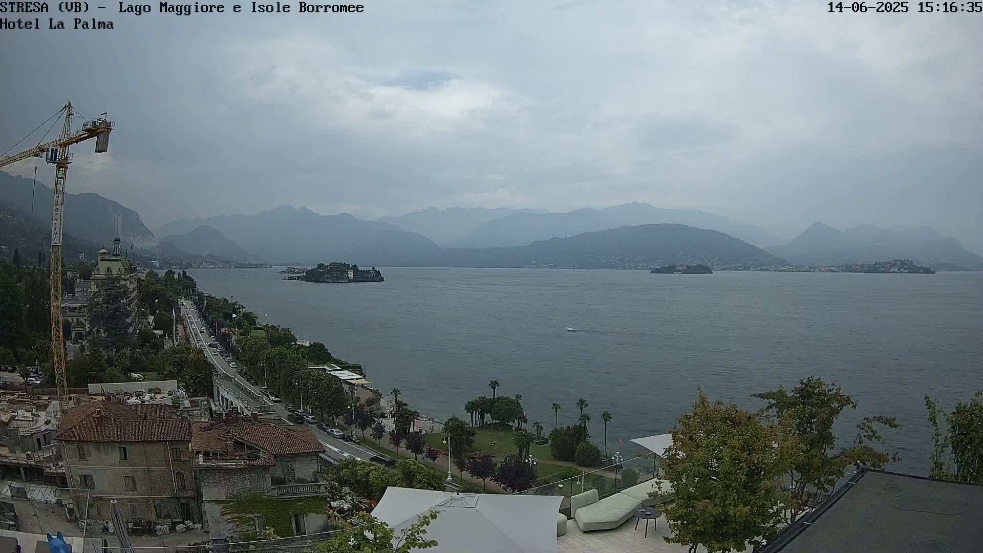 A view of a lake with distant mountains under a cloudy sky, showing a shoreline with buildings, trees, and a construction crane.