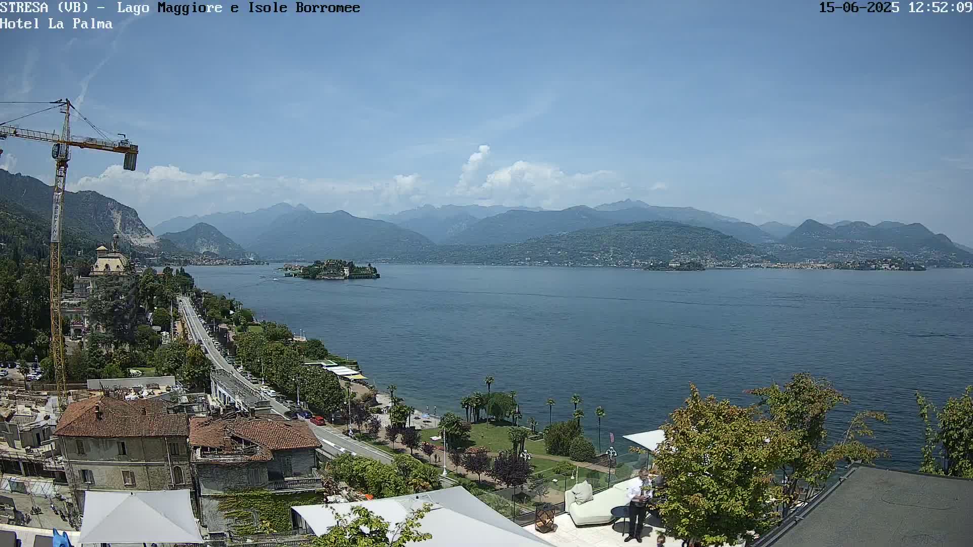 A partly cloudy day overlooking a large lake with mountains in the distance, featuring a shoreline with buildings, trees, and a construction crane.