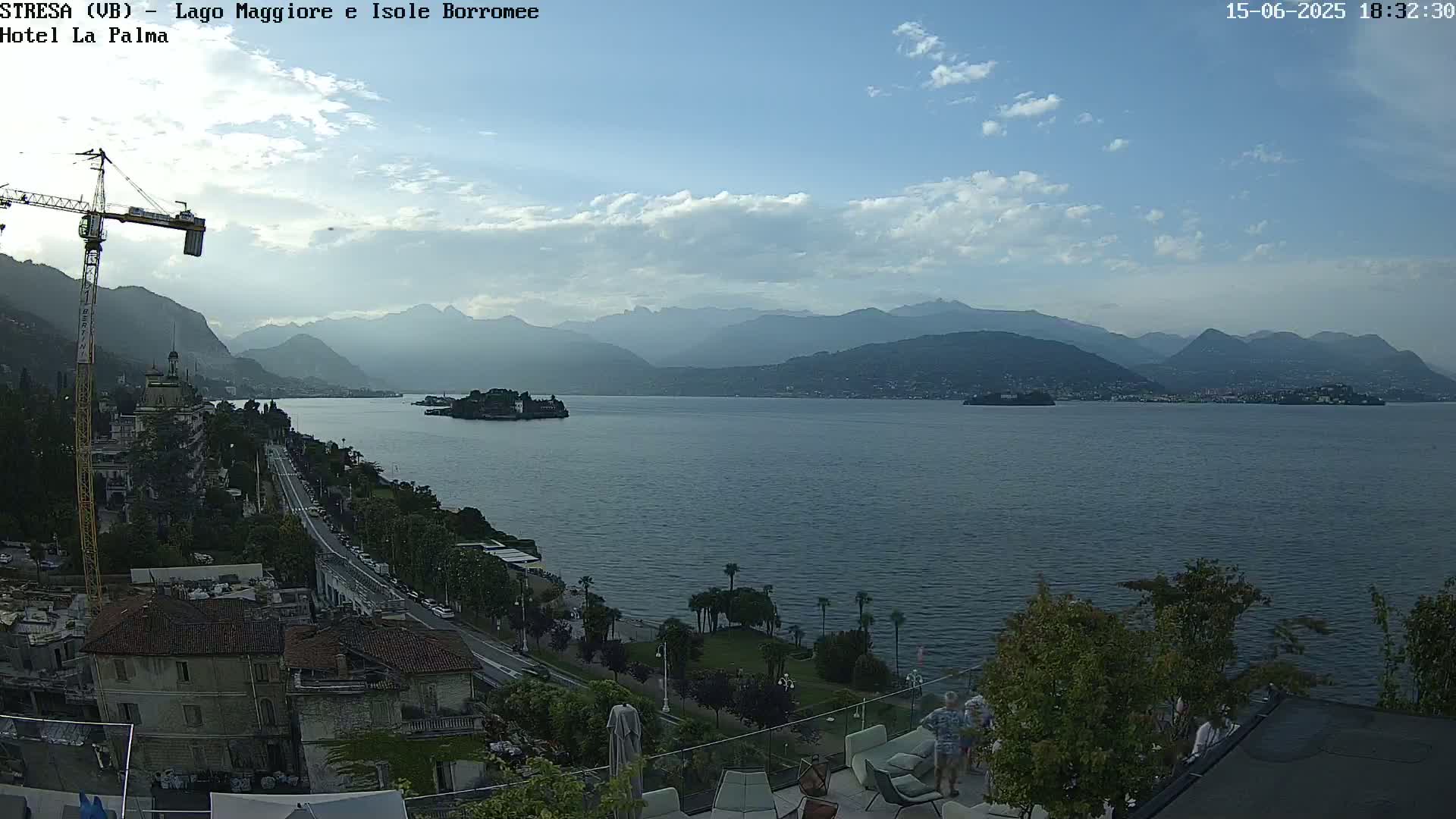 A partly cloudy day overlooking a large lake with islands and distant mountains, showing a town with a construction crane and people relaxing on an outdoor patio.