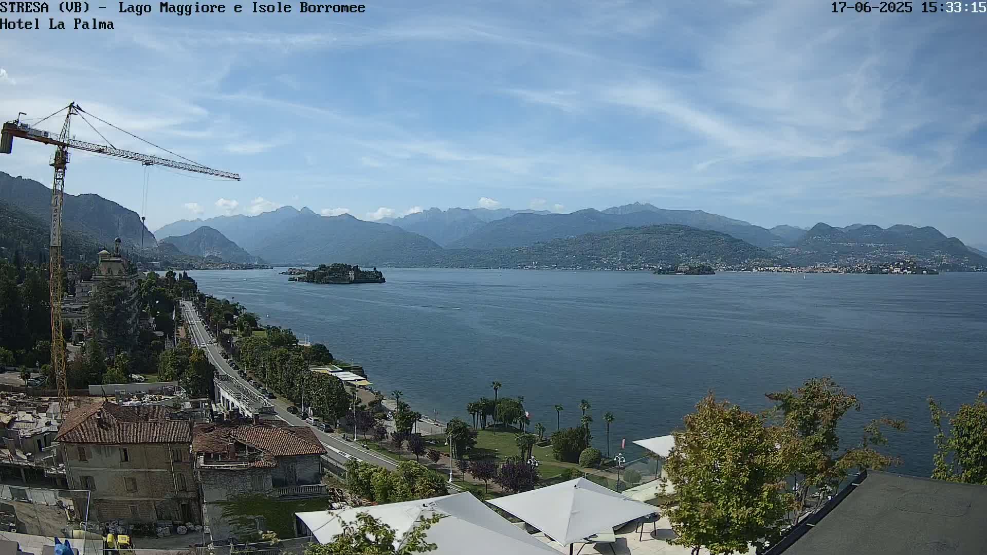 A partly cloudy day reveals a large lake surrounded by mountains, with a town, a construction crane, and palm trees visible in the foreground.