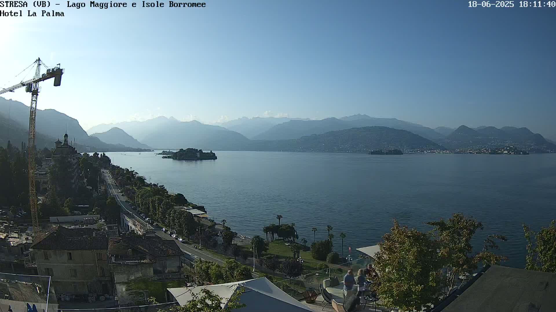 A clear, sunny day overlooking a large lake with mountains in the distance, a small island in the lake, and a town with a construction crane visible in the foreground.