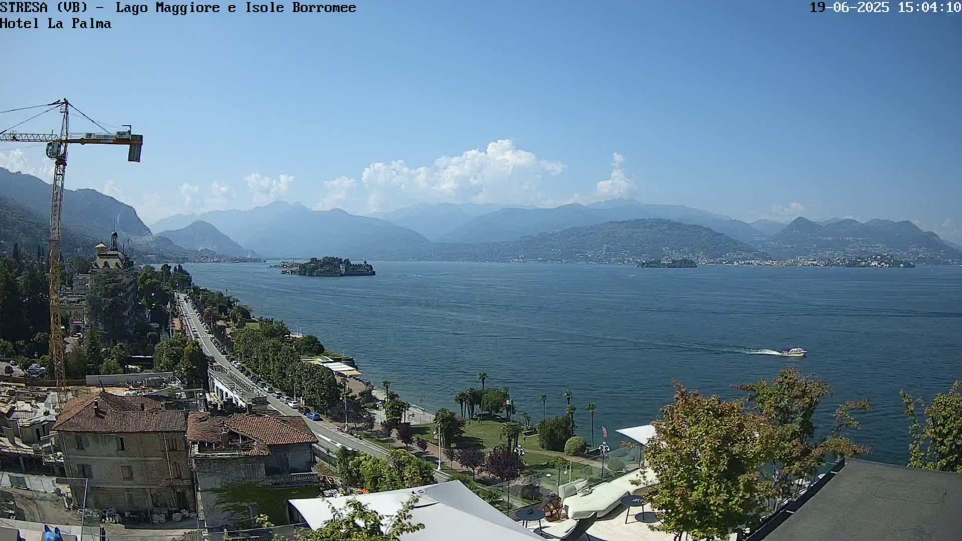 A partly cloudy sunny day overlooks a large lake with mountains in the distance, a town along the shoreline, and a small boat on the water.