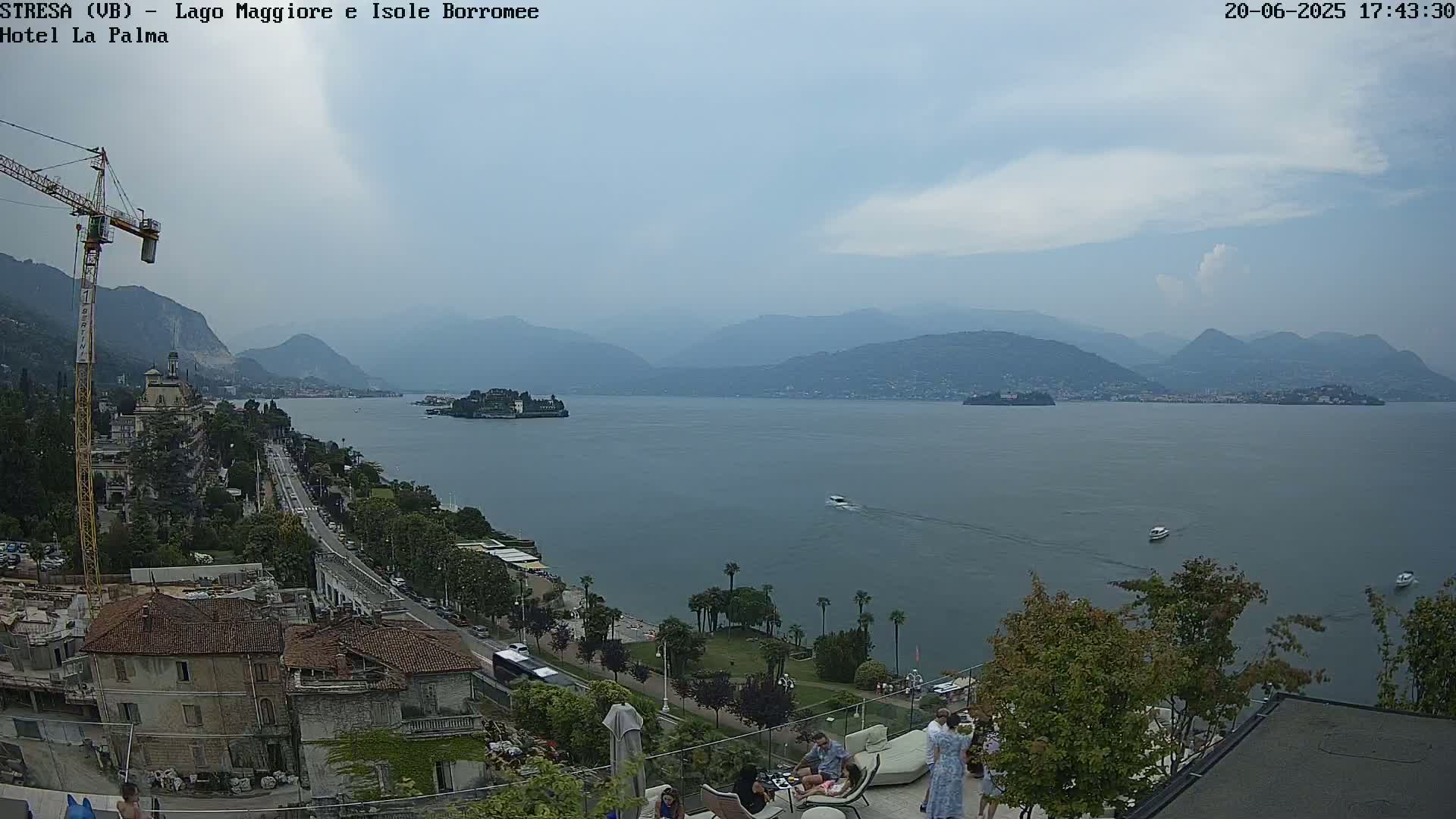 An elevated view of a lake with mountains in the background, under a mostly cloudy sky; a town is visible on the lakeshore, with boats on the water and people gathered near a building.