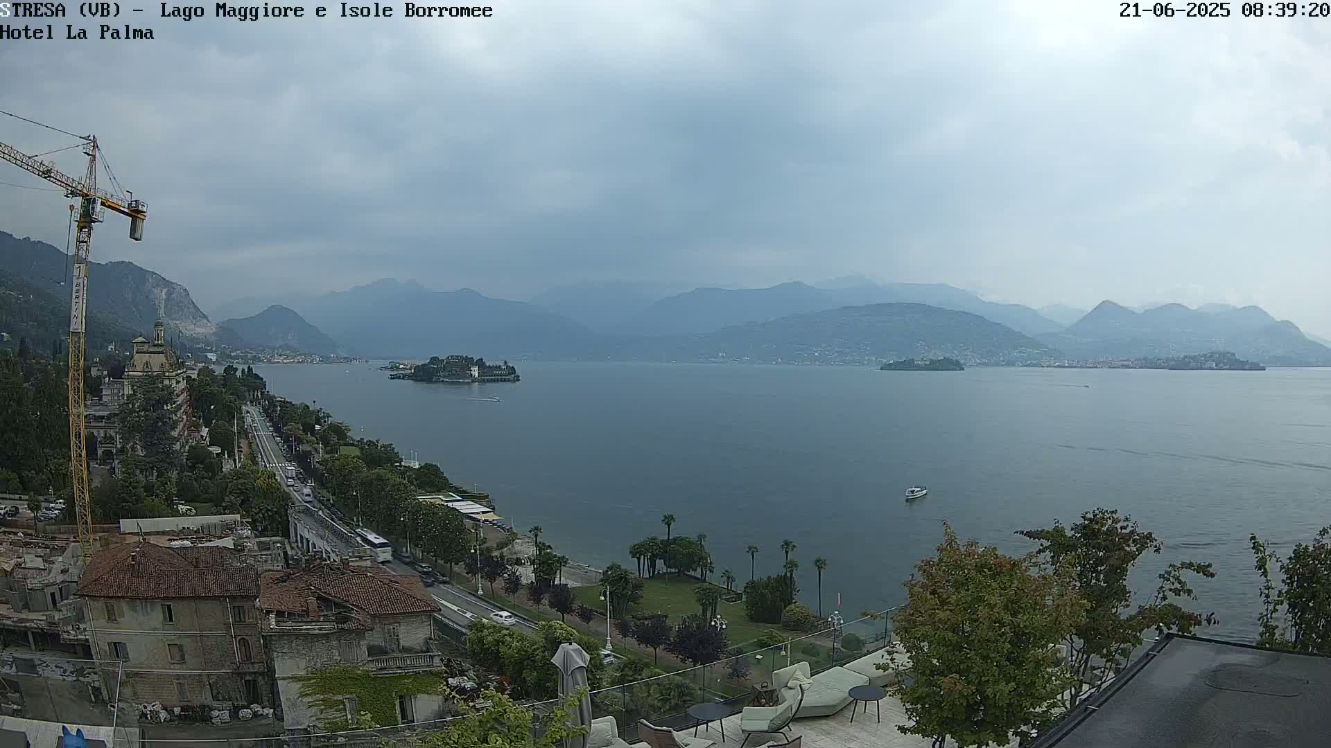 A view of a large lake with mountains in the background under an overcast sky, showing a town and a construction crane on the left and palm trees on the right along the shore.