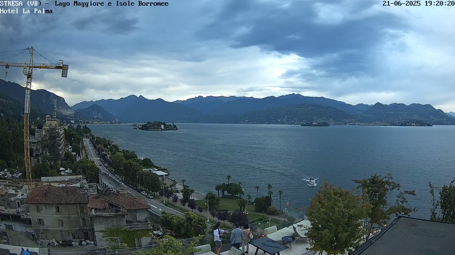 A partly cloudy day reveals a lake view, with mountains in the background, a small island in the foreground, and people gathered on a patio overlooking the water.