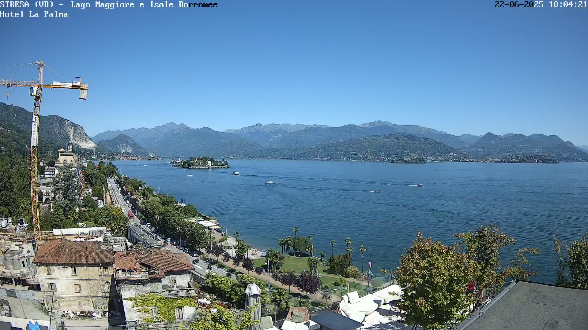A sunny view of a lake with mountains in the background, a shoreline with buildings and vegetation, and several boats on the water.
