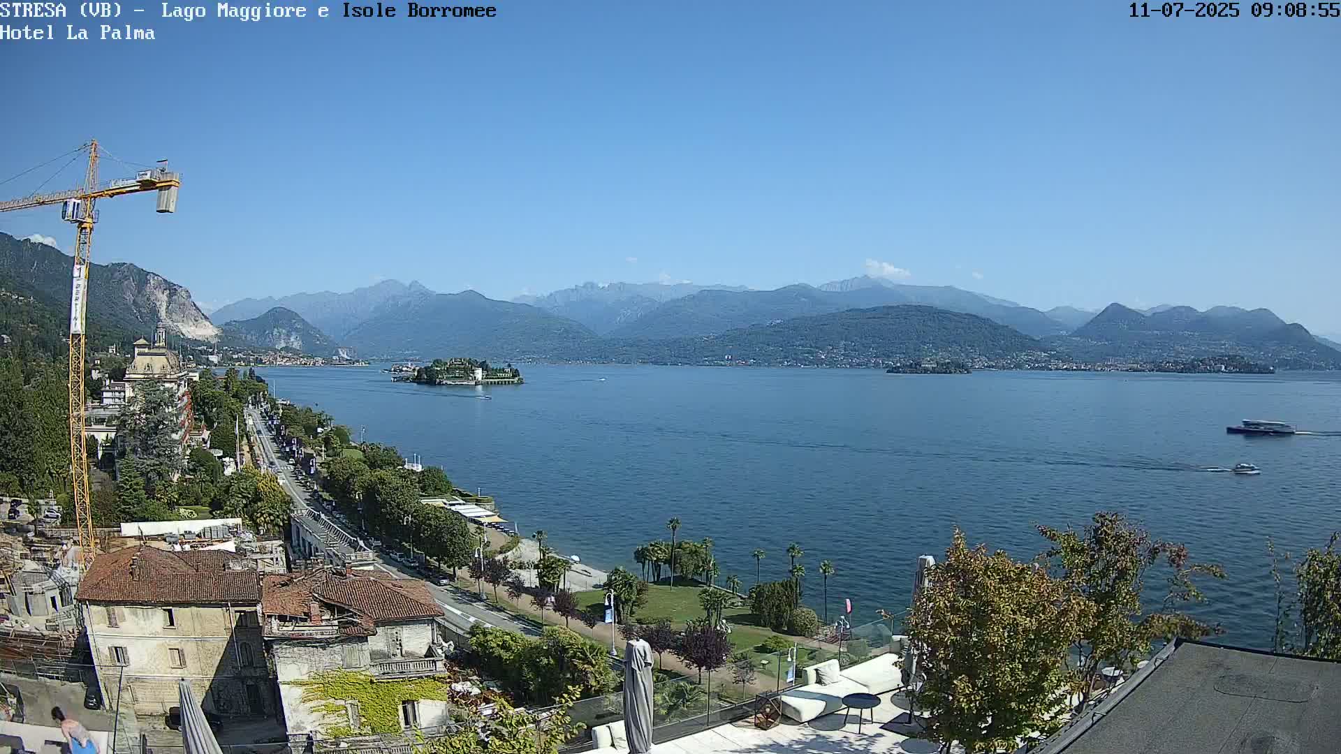 A sunny day overlooking a large lake with mountains in the background, featuring buildings, trees, and boats on the water.
