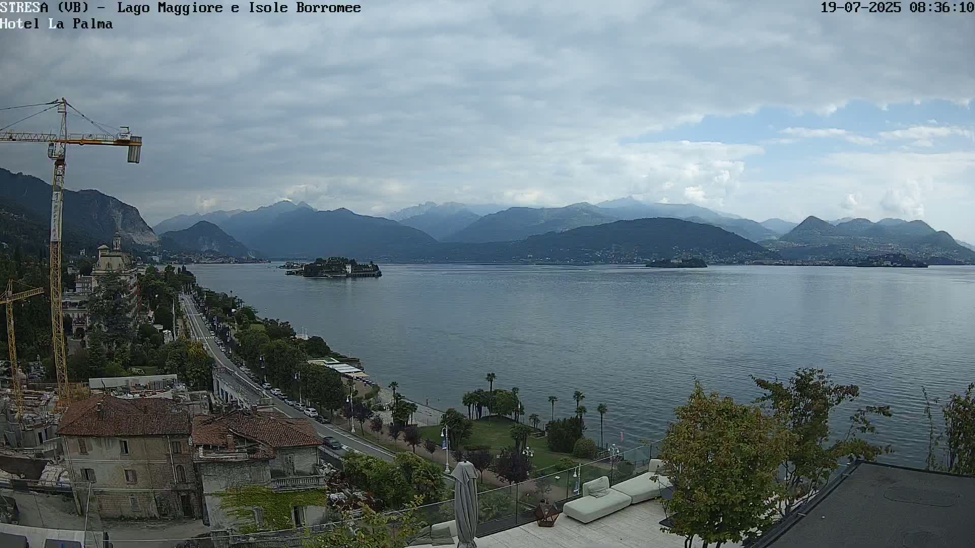 A partly cloudy day reveals a lake view with mountains in the distance, a shoreline with buildings and trees, and a construction crane.