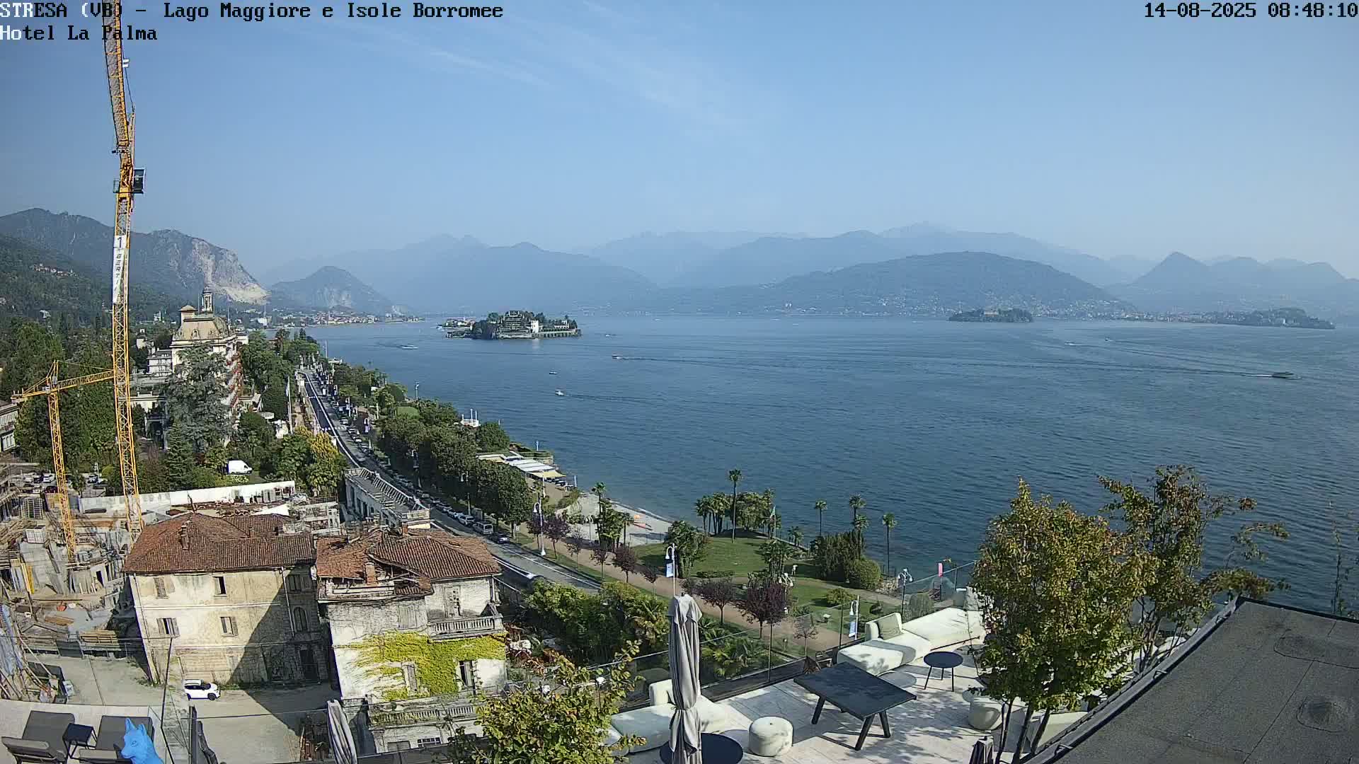 A sunny, high-angle view shows a large lake with islands and distant mountains, a shoreline with buildings, palm trees and a road, and a rooftop patio with furniture in the foreground.