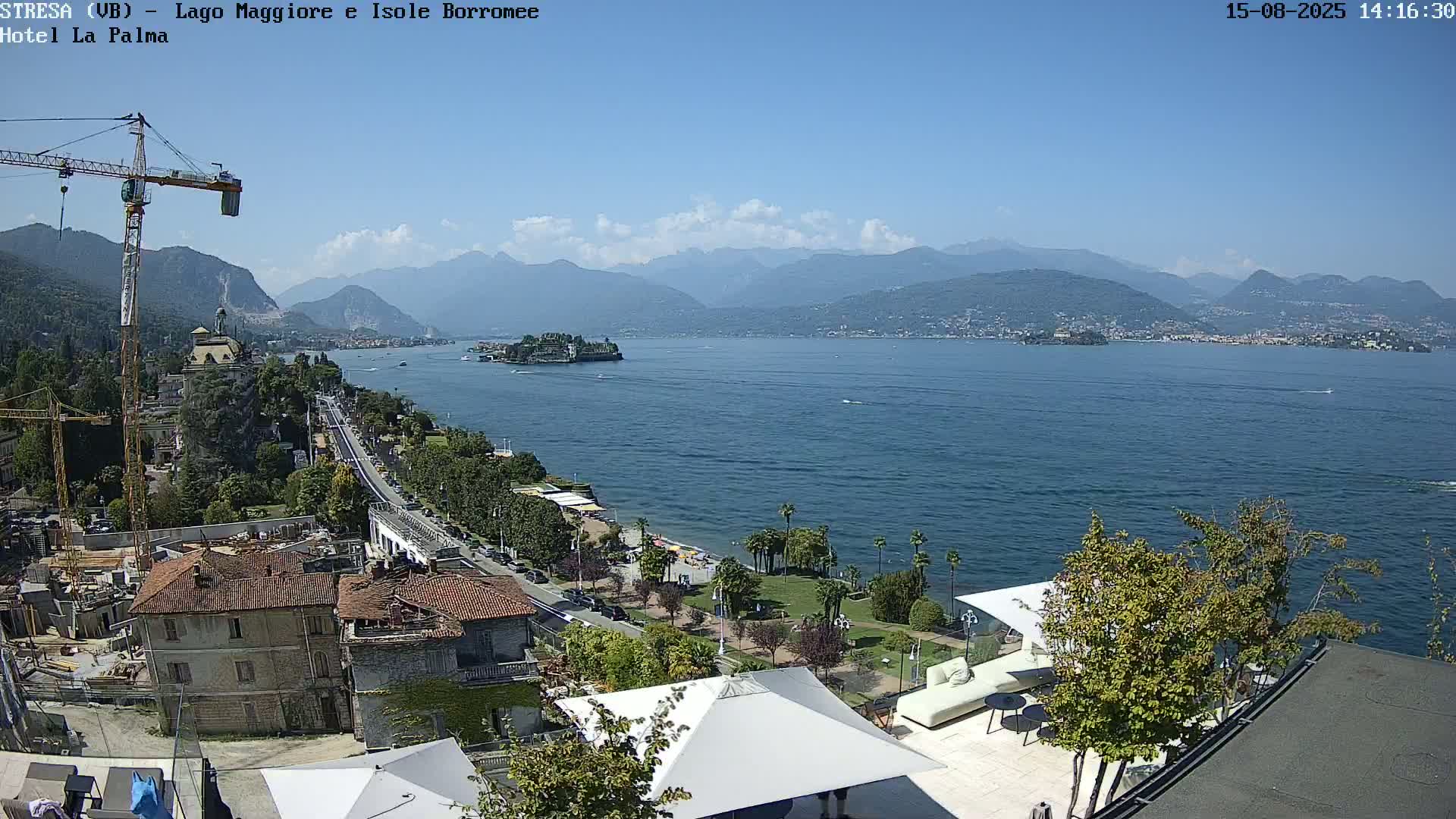 A sunny, high-angle view shows a large lake with islands and distant mountains, a shoreline with buildings, palm trees and a road, and a rooftop patio with furniture in the foreground.