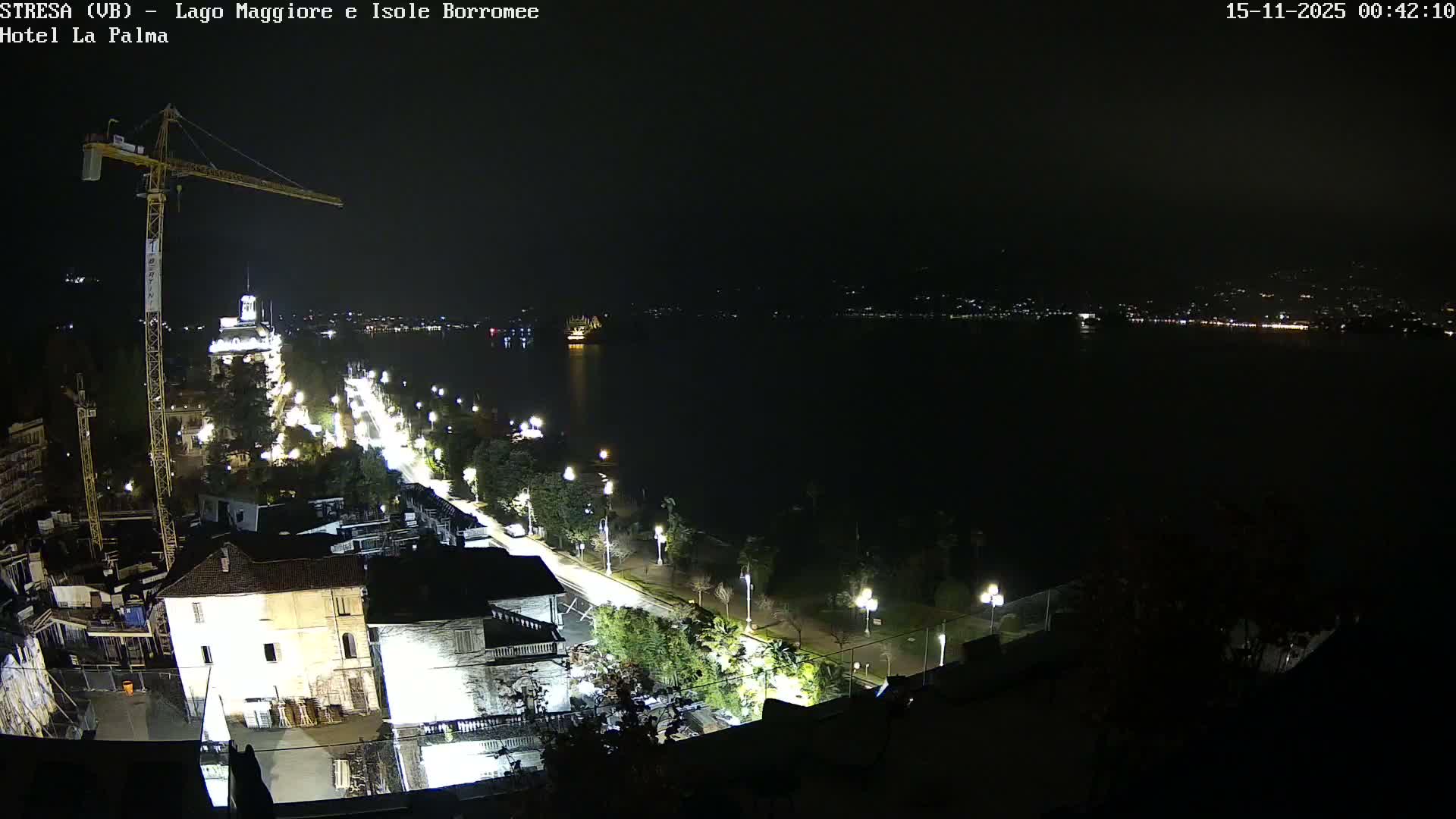 The image displays a clear night over a lakeside town, featuring a brightly lit promenade, buildings, and a large construction crane in the foreground, with distant city lights reflecting across the calm water.