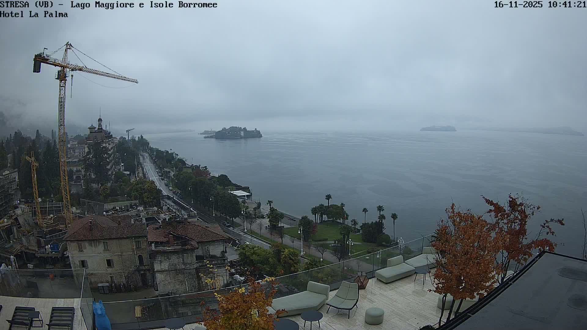 An elevated view reveals a lakeside town with a prominent construction crane and a curving road, several islands dimly visible across the vast, calm water under an overcast and misty sky, with a modern rooftop terrace in the foreground.