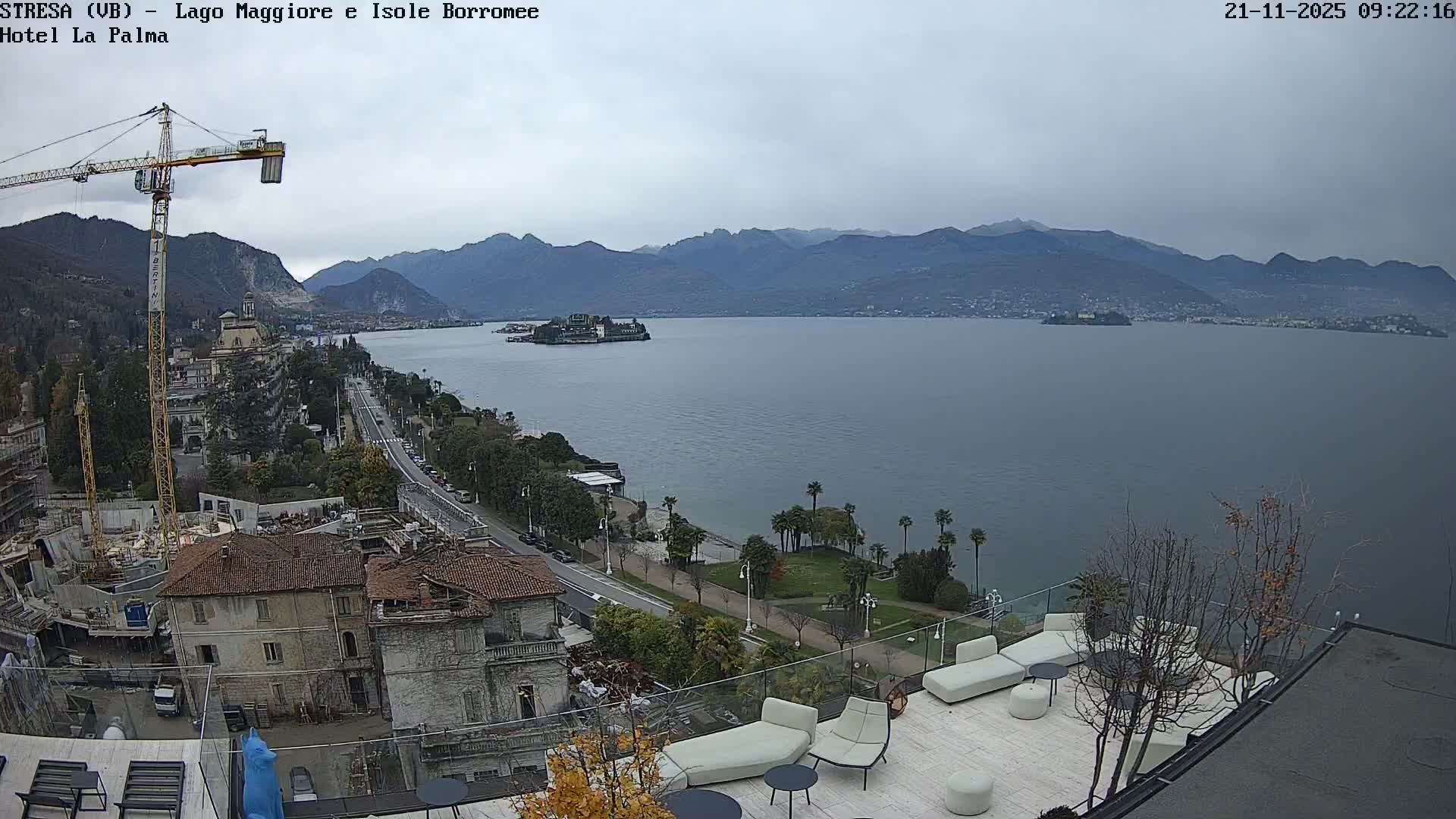 On an overcast day, an elevated perspective reveals a modern rooftop patio and a construction site in the foreground, looking out over a busy lakeside road, a wide lake with multiple islands, and distant mountains under a cloudy sky.