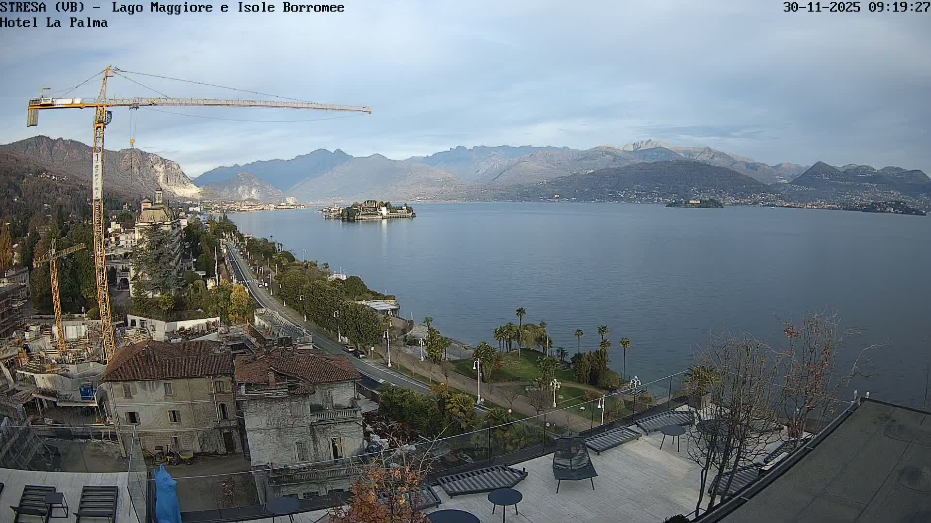 On a clear day with scattered clouds, a wide shot reveals a picturesque lakeside town with a prominent construction crane, a busy road along the water, an island in the expansive lake, and a backdrop of numerous mountains.