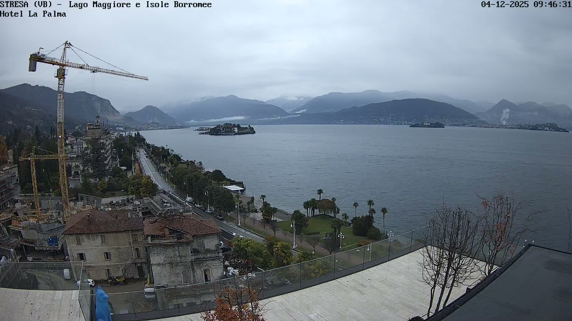 Under an overcast sky, a large lake with distant hazy mountains and an island featuring buildings is overlooked by a scenic coastline with a road, various structures, and a prominent construction crane in the foreground.