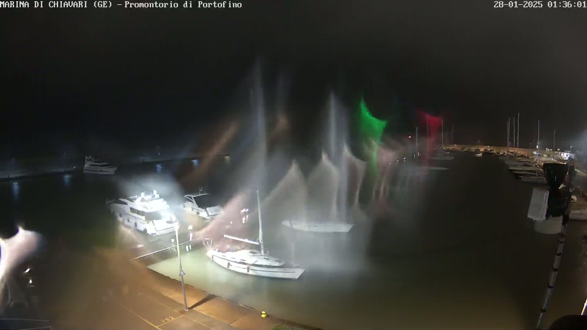 Several boats are moored in a harbor at night during a storm with heavy spray obscuring visibility.