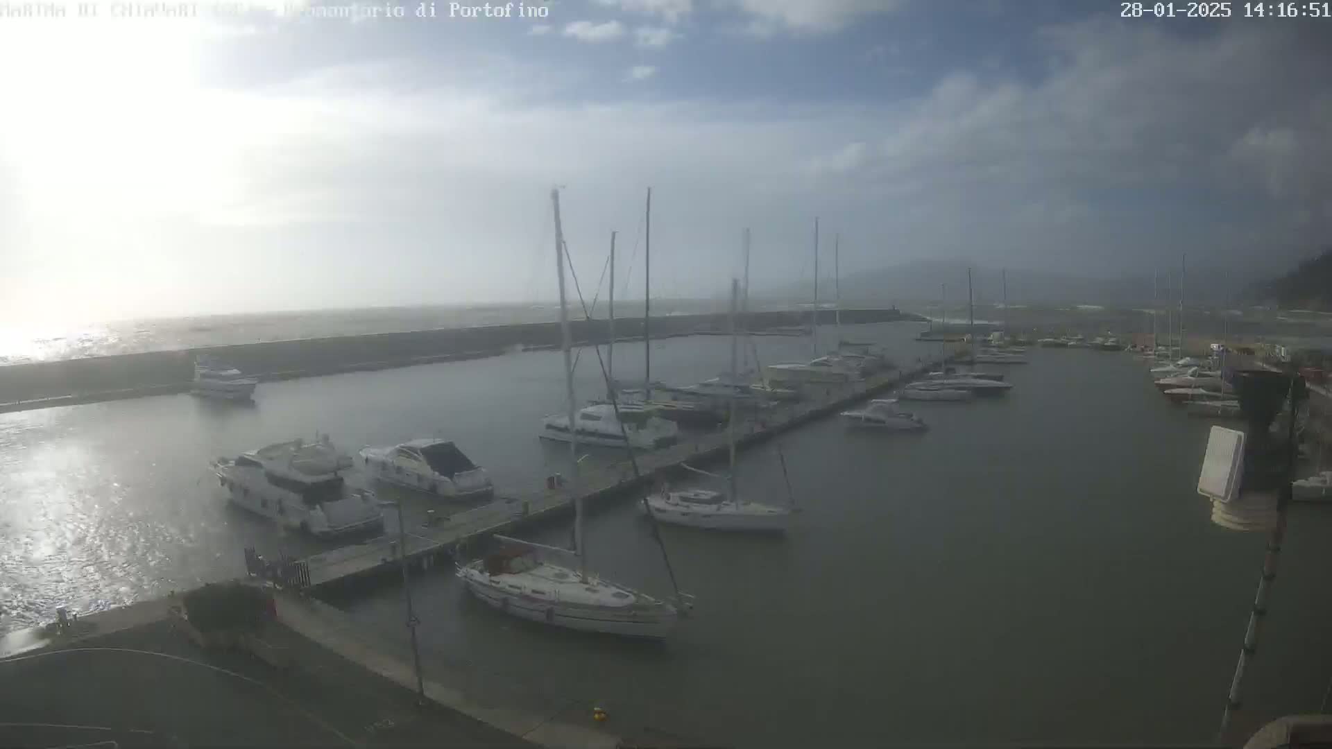 A marina filled with sailboats and motorboats is partially sheltered by a seawall under an overcast sky with choppy water.