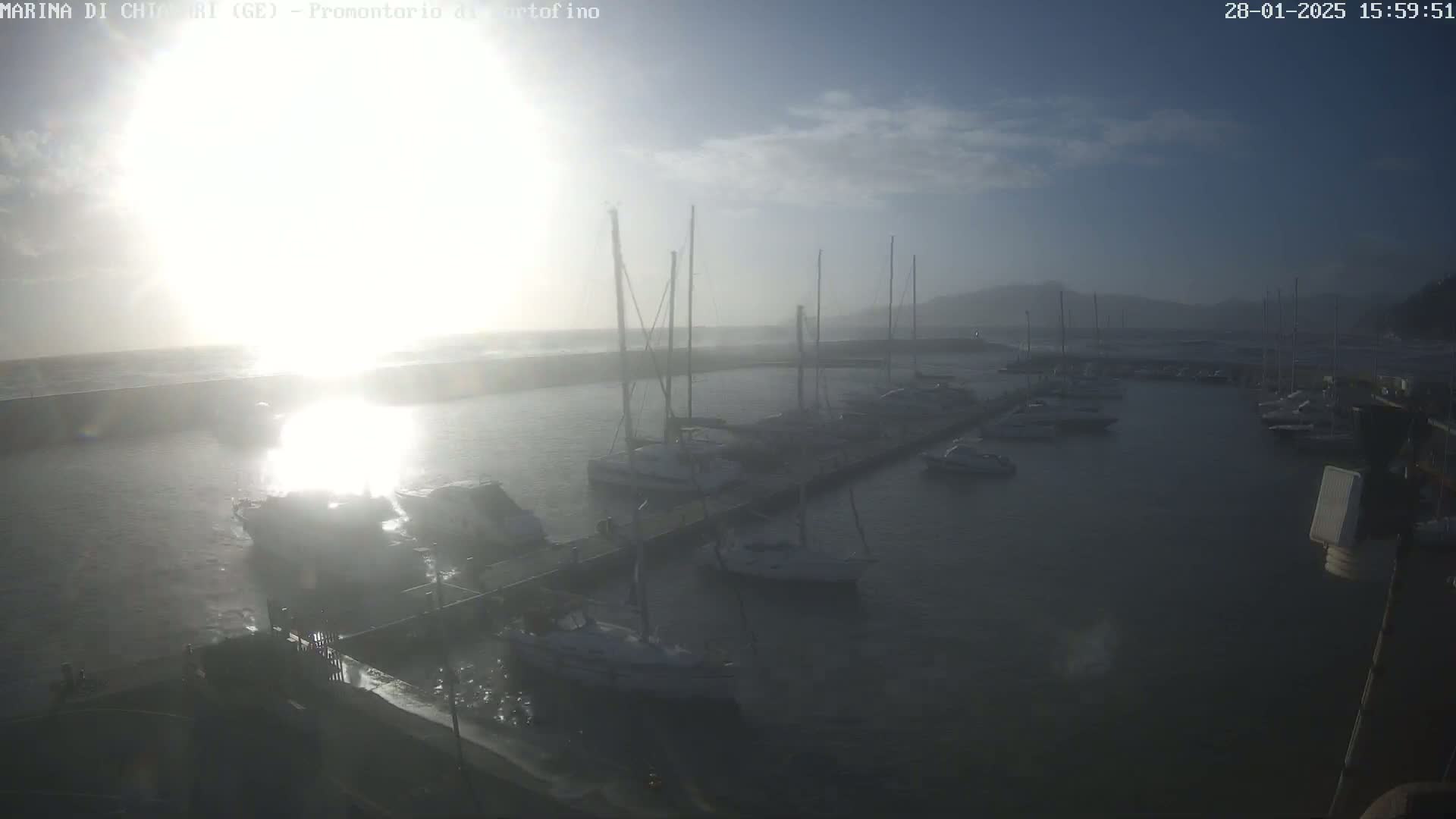 A hazy, sunny day shows a marina filled with sailboats and motorboats docked along a pier, with choppy water visible beyond the harbor wall.