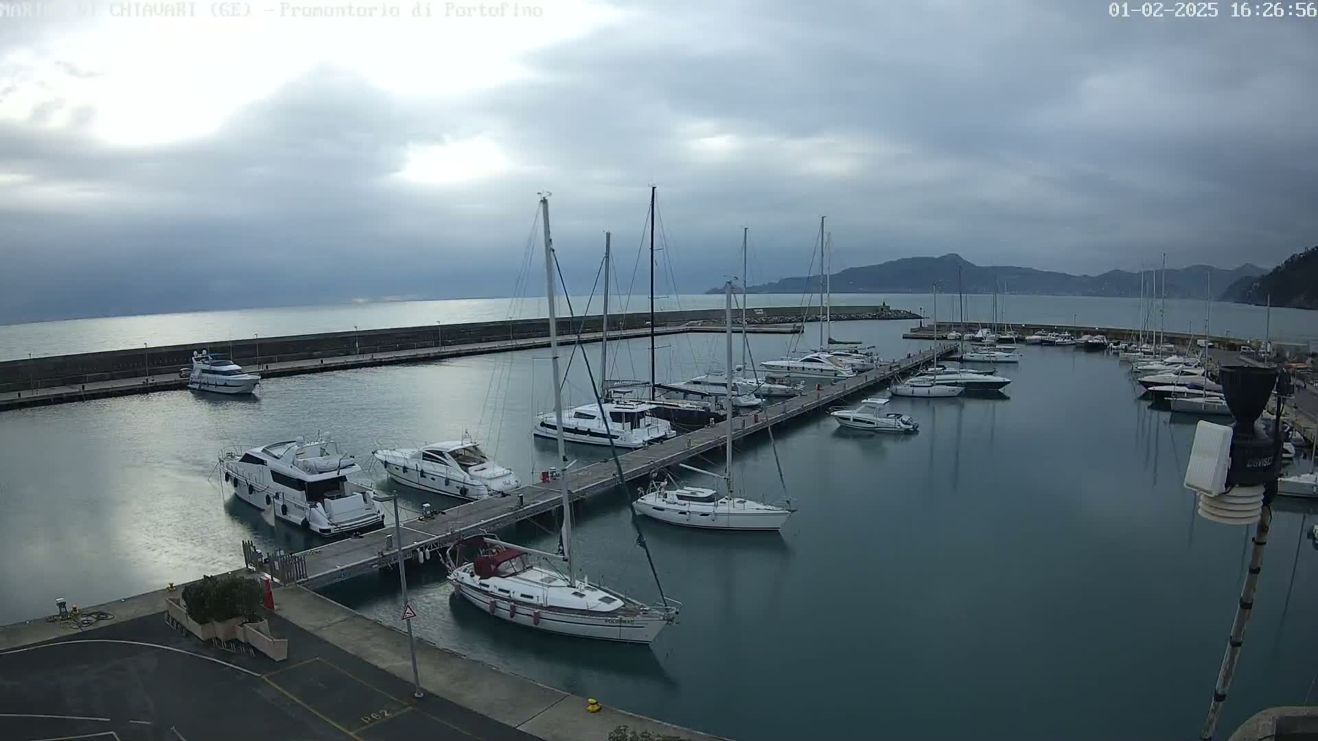 A marina filled with sailboats and motorboats on a cloudy day, with calm water and distant hills visible in the background.
