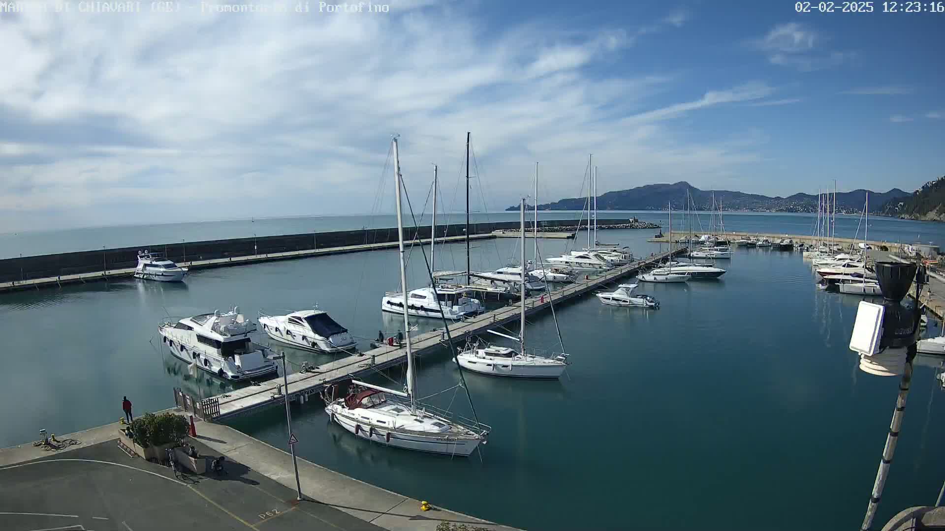 A marina filled with sailboats and motorboats is nestled against a seawall under a mostly sunny sky with a few clouds.