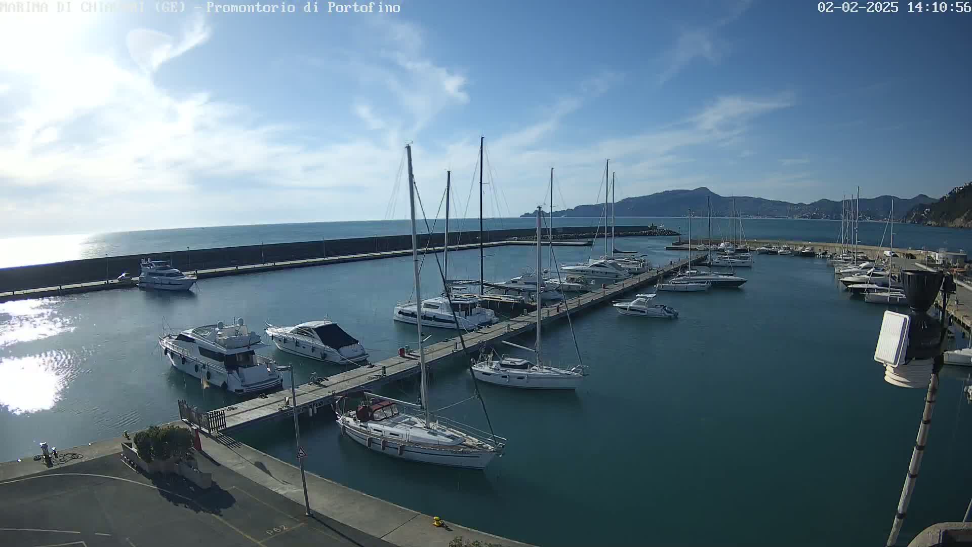 A marina filled with sailboats and motorboats is nestled in a calm harbor under a mostly sunny sky, with mountains visible in the distance.