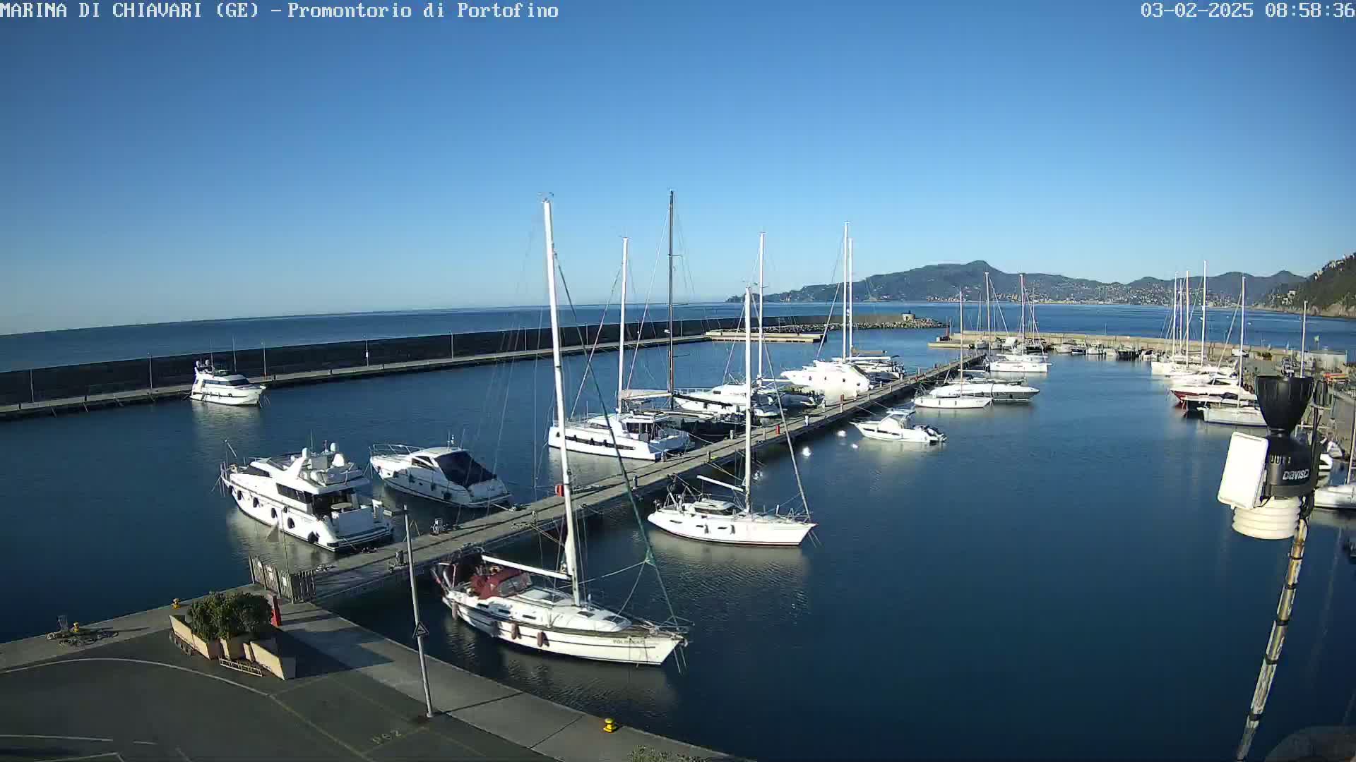 A marina filled with sailboats and motorboats on a calm, sunny day, with mountains visible in the distance.