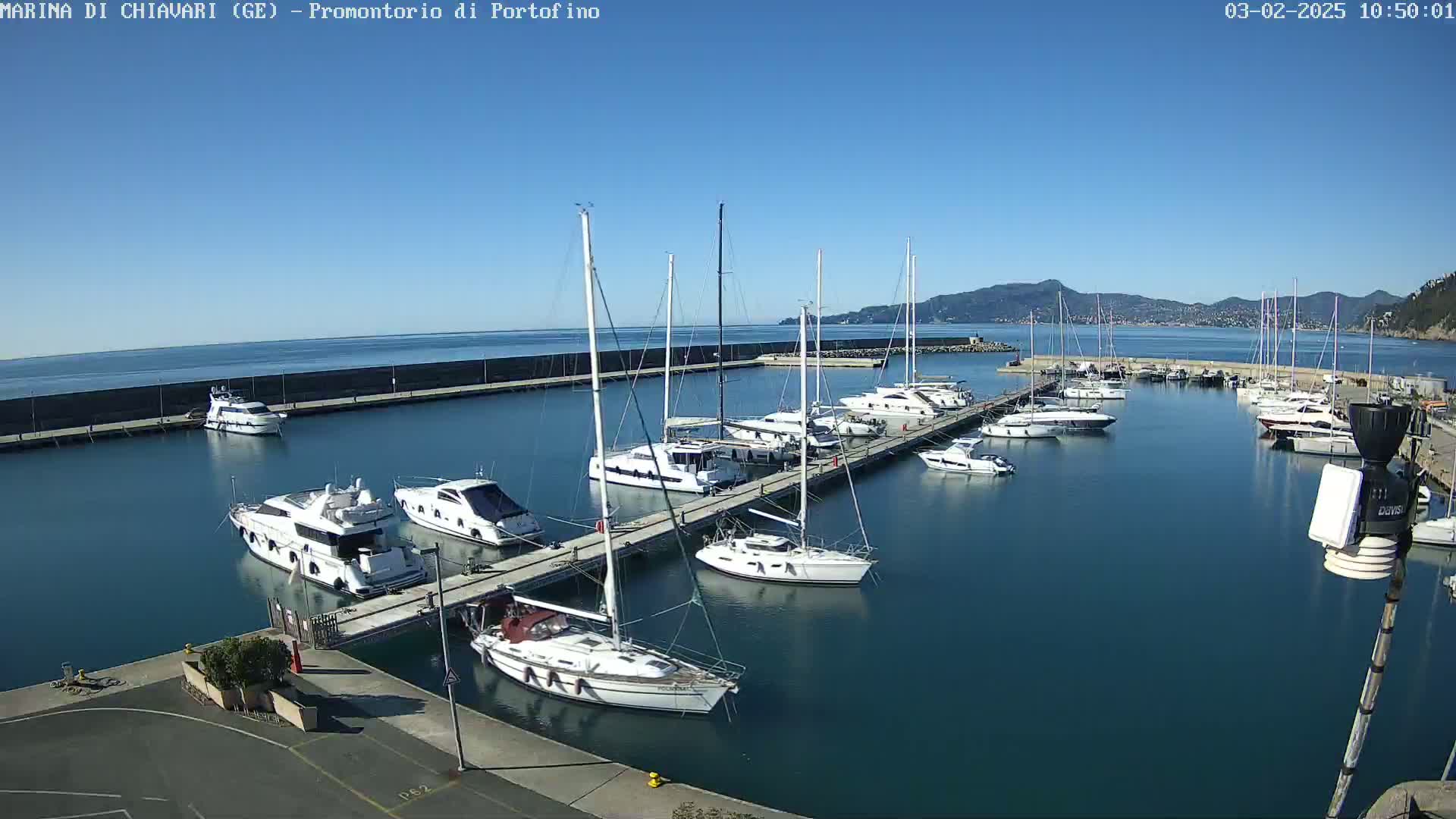 A marina filled with sailboats and motorboats on a sunny day, with calm blue water and distant mountains visible.