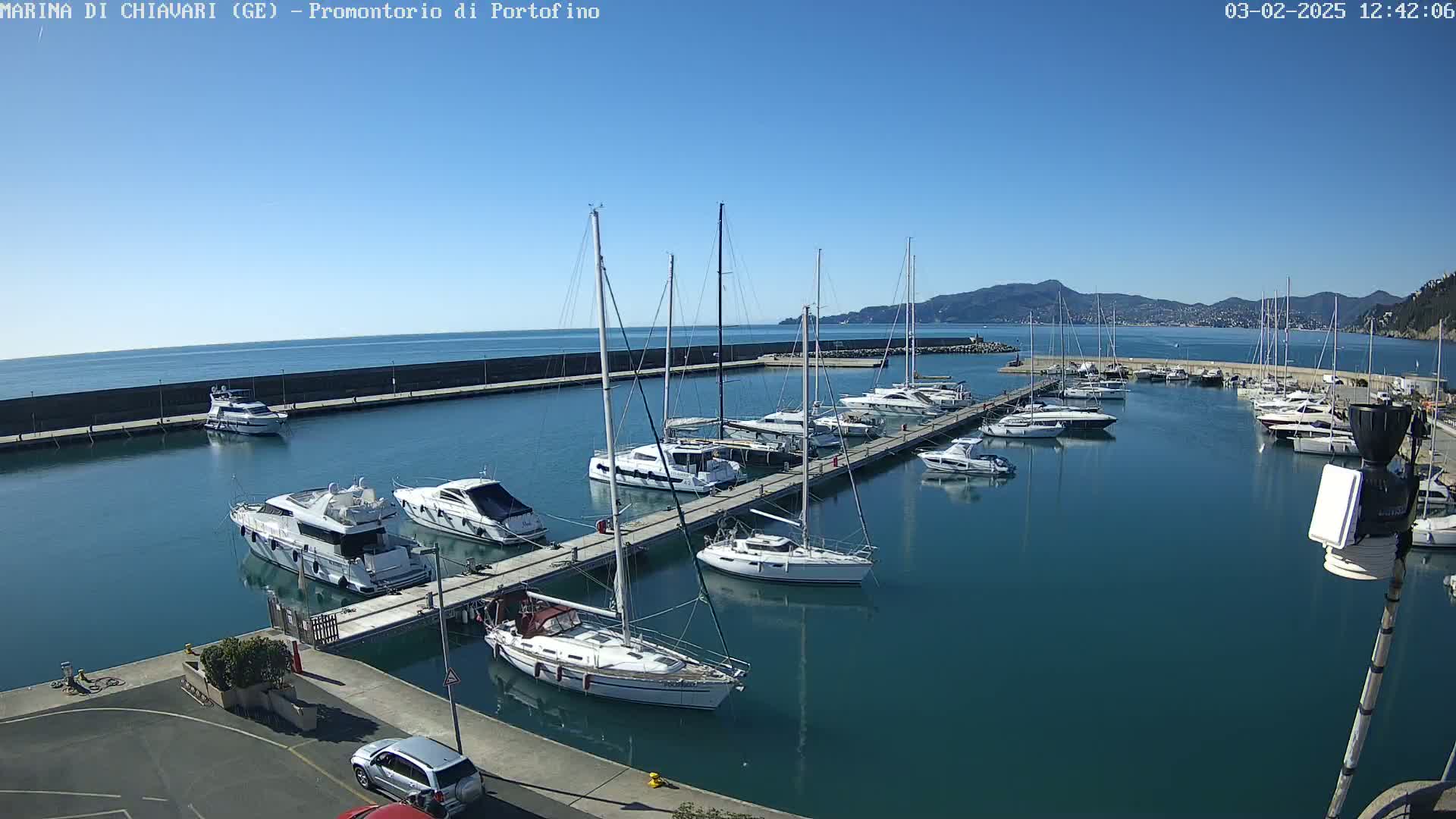 A marina filled with numerous sailboats and motor yachts on a sunny day, with calm waters and a mountain range visible in the background.