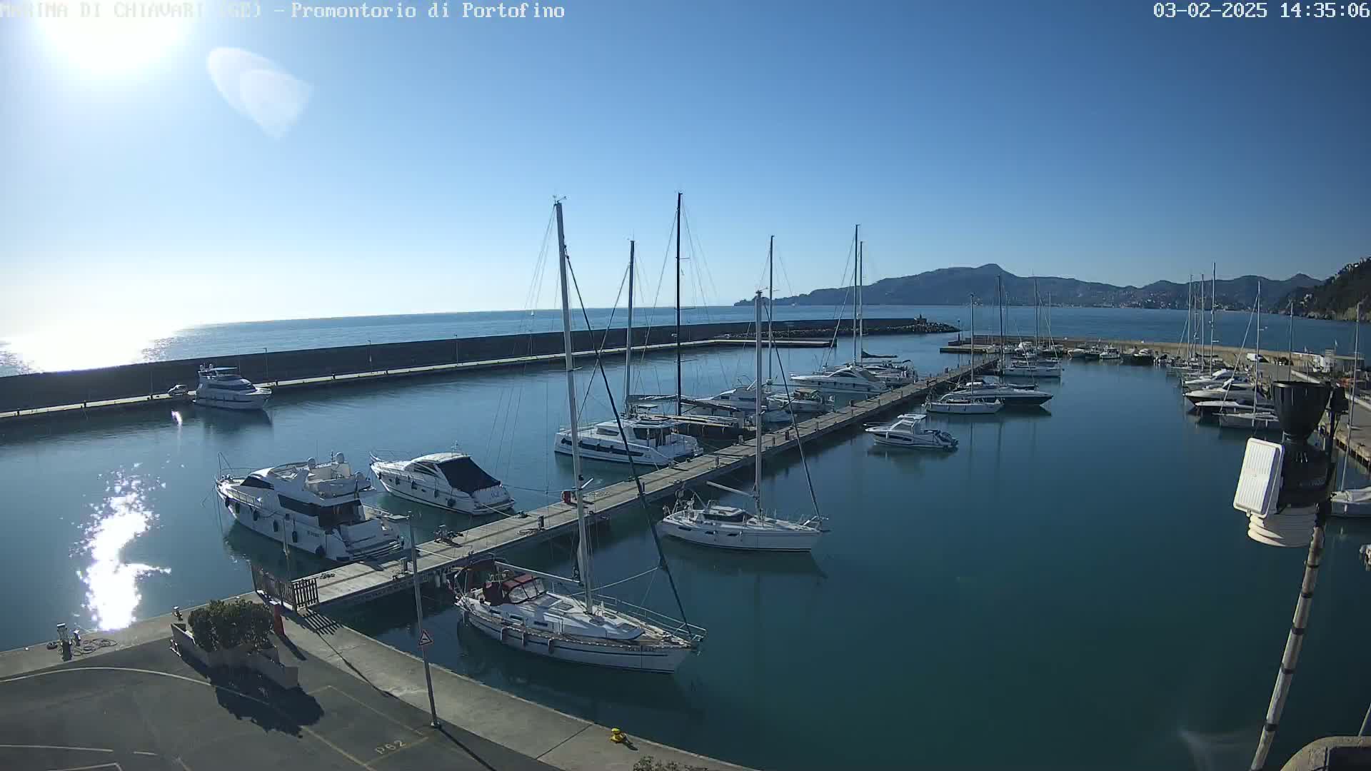 A marina filled with sailboats and motorboats is situated on calm, clear blue water under a sunny sky, with hills visible in the background.