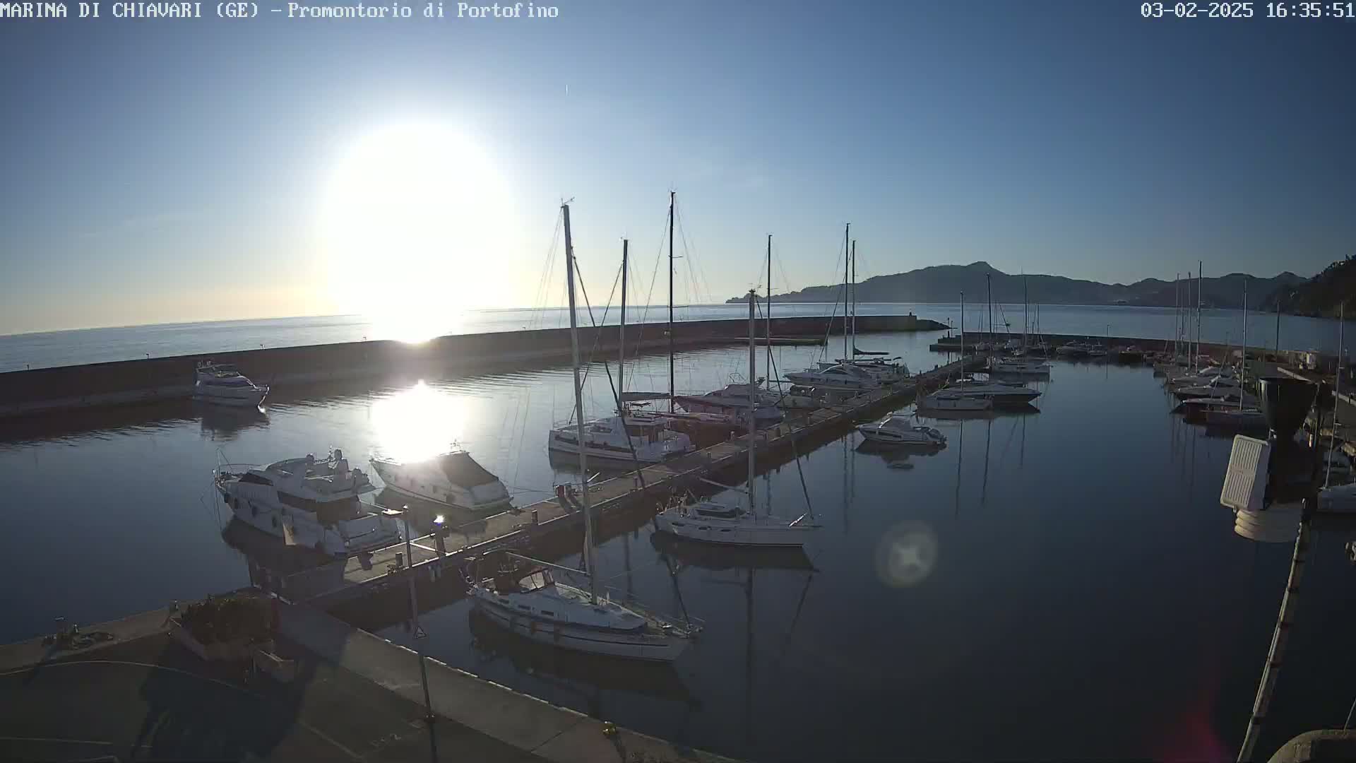 A marina filled with sailboats and motorboats is seen at sunrise, with calm water reflecting the sunlight and a distant mountain range visible.