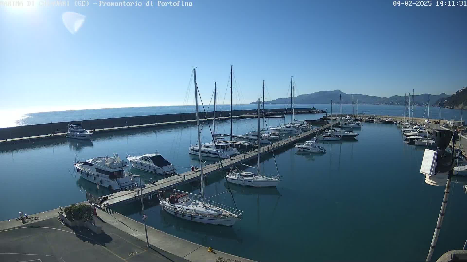 A marina filled with sailboats and motorboats is situated on calm, clear water under a bright, sunny sky, with mountains visible in the distance.