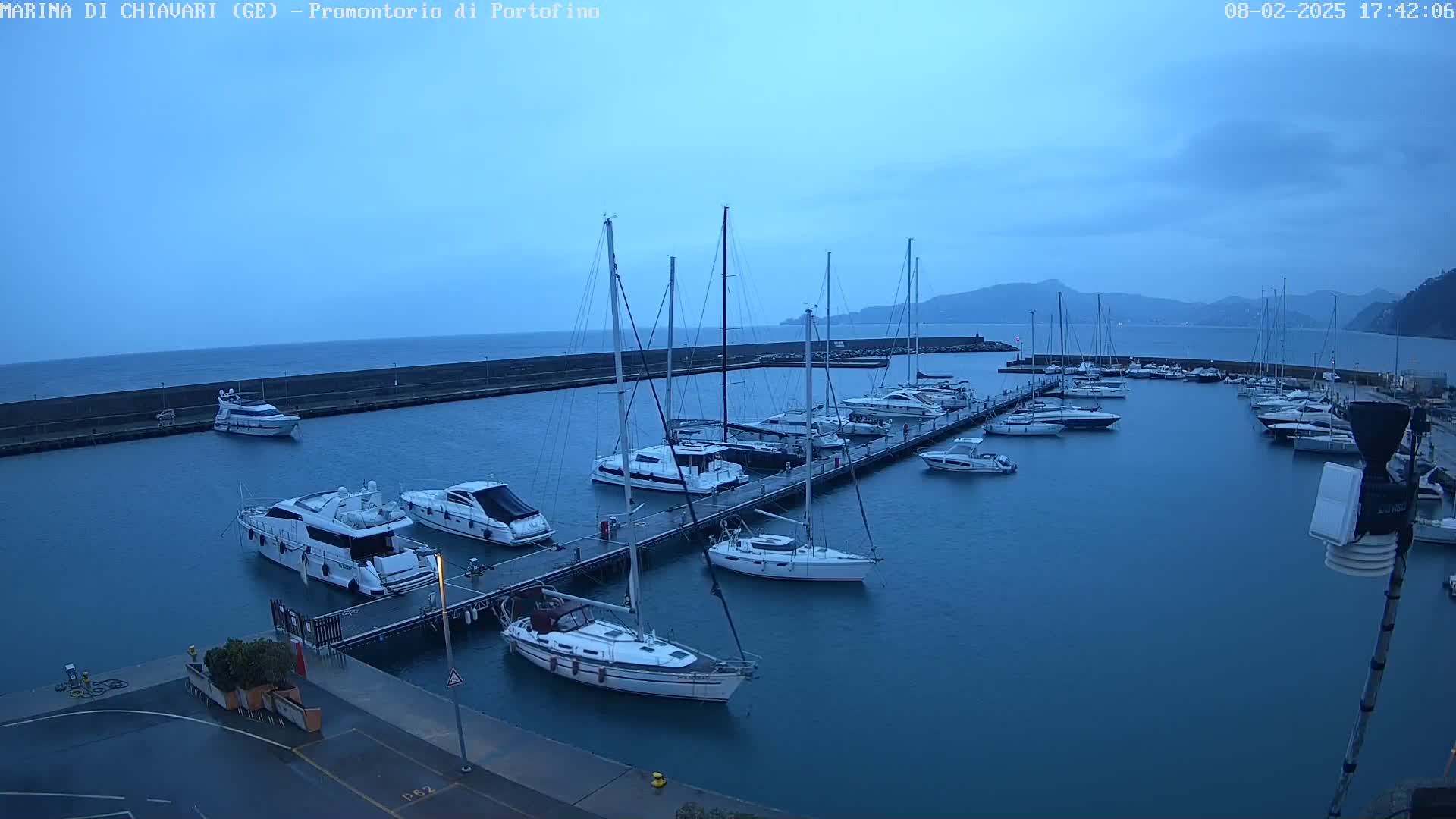 Numerous yachts and sailboats are moored at a marina on a calm, dusky blue sea, with hills visible in the background under an overcast sky.