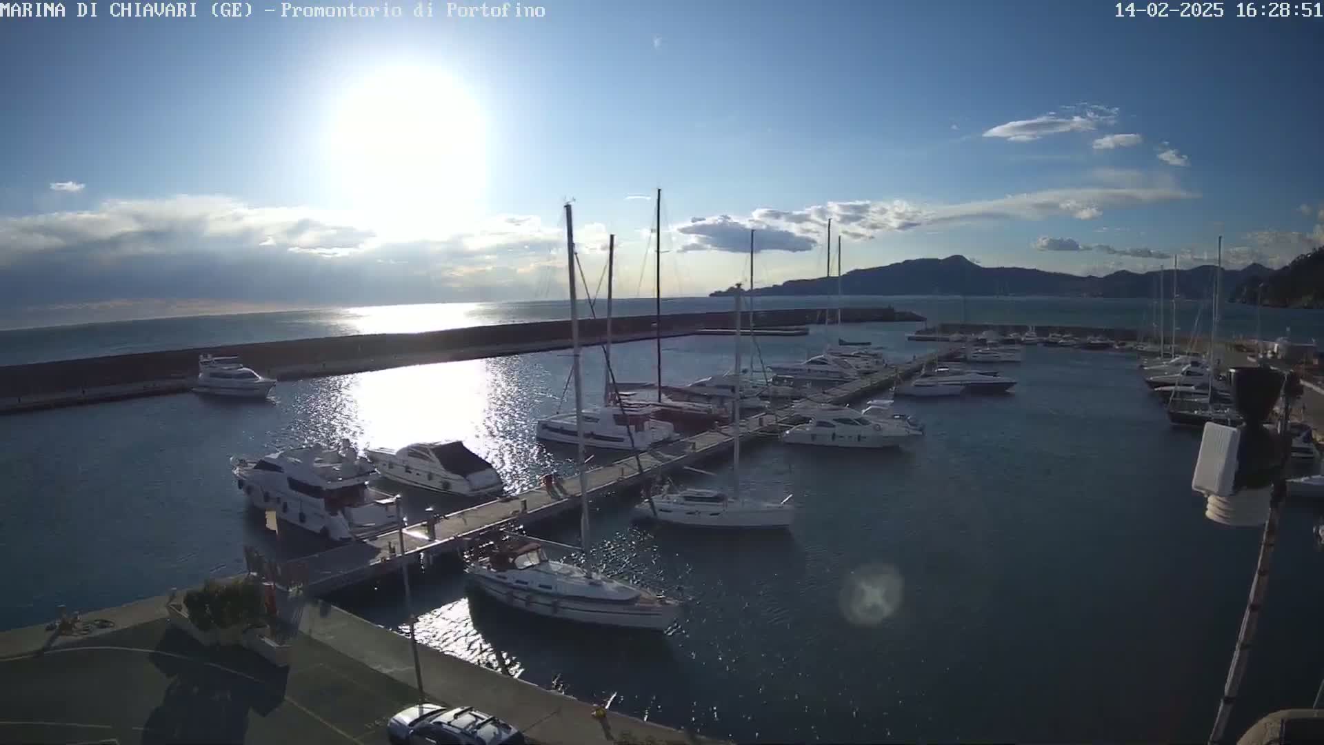 A marina filled with sailboats and motorboats is seen under a mostly sunny sky, with a few clouds and mountains in the distance.