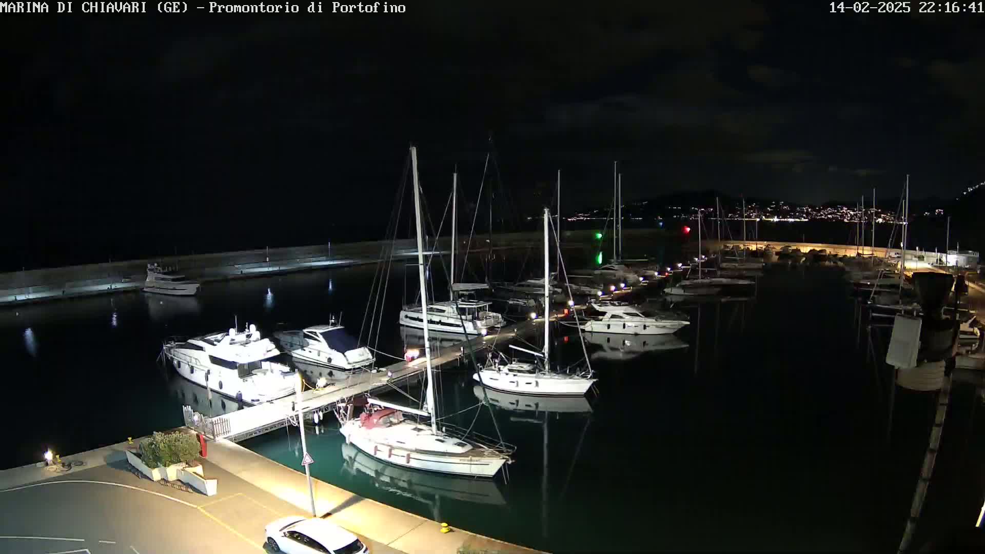 A nighttime aerial view shows a marina filled with sailboats and motor yachts, docked along a seawall under a dark sky.
