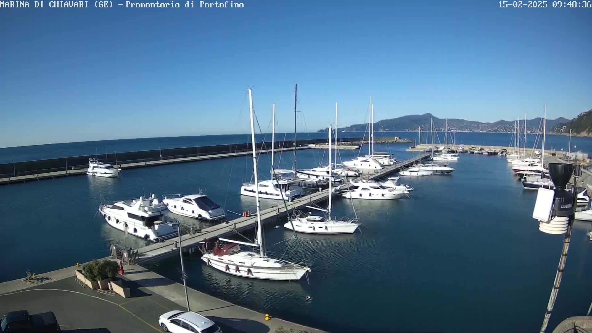 A marina filled with sailboats and motorboats on a sunny day, with mountains visible in the distance.