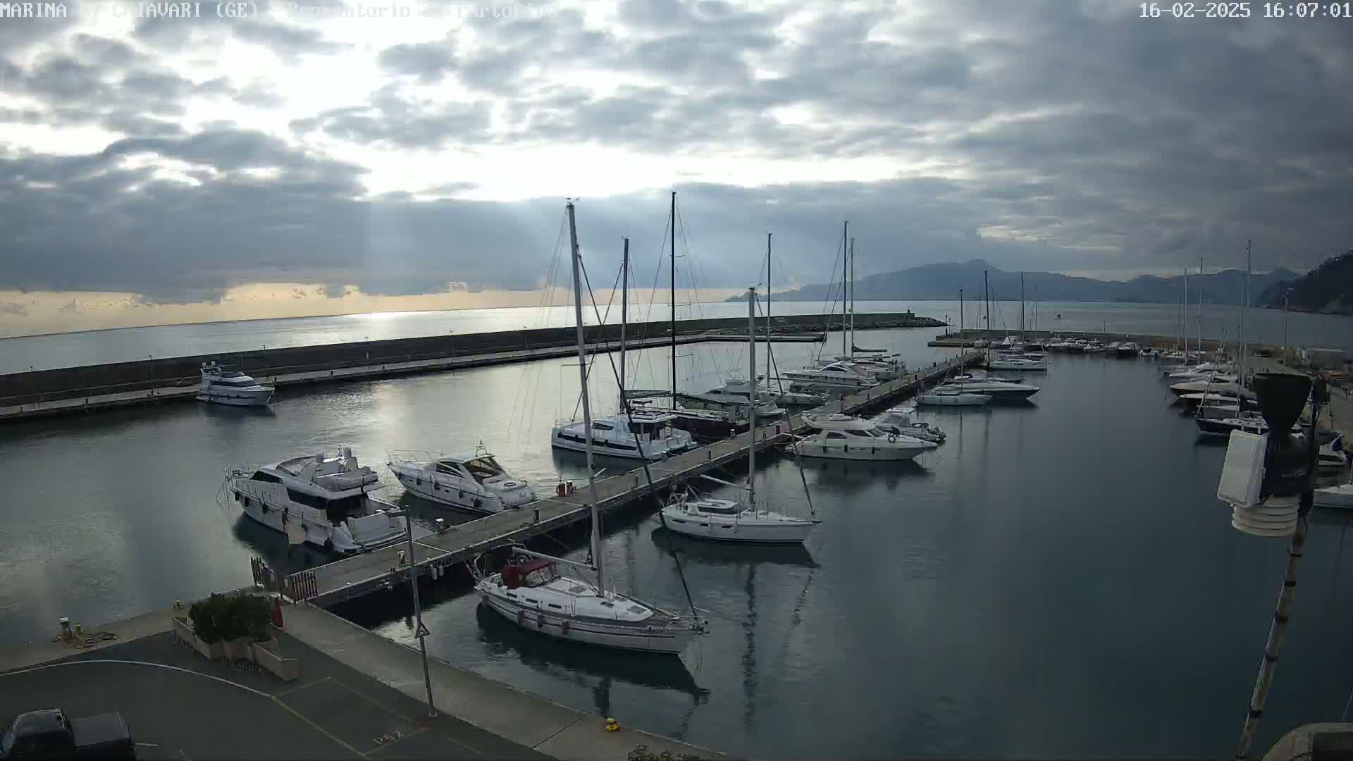 Many boats are docked at a marina on a partly cloudy day, with mountains visible in the distance.